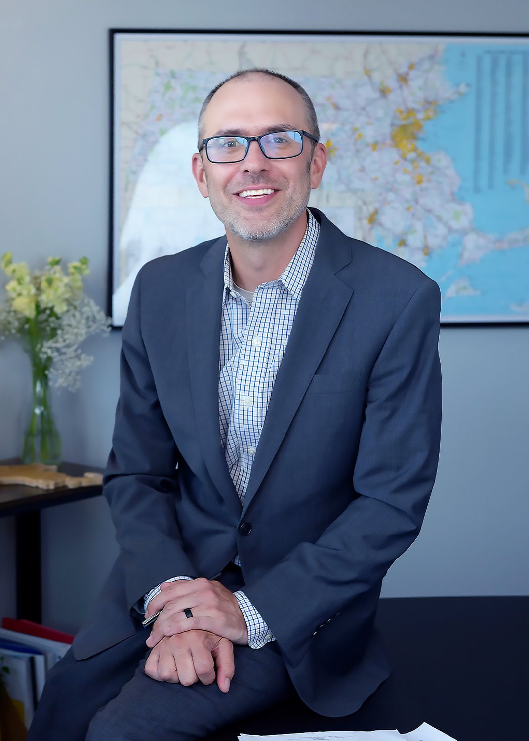 a  smiling man wearing a blue suit, glasses, and a checkered shirt sitting in an office, smiling at the camera with a map in the background.