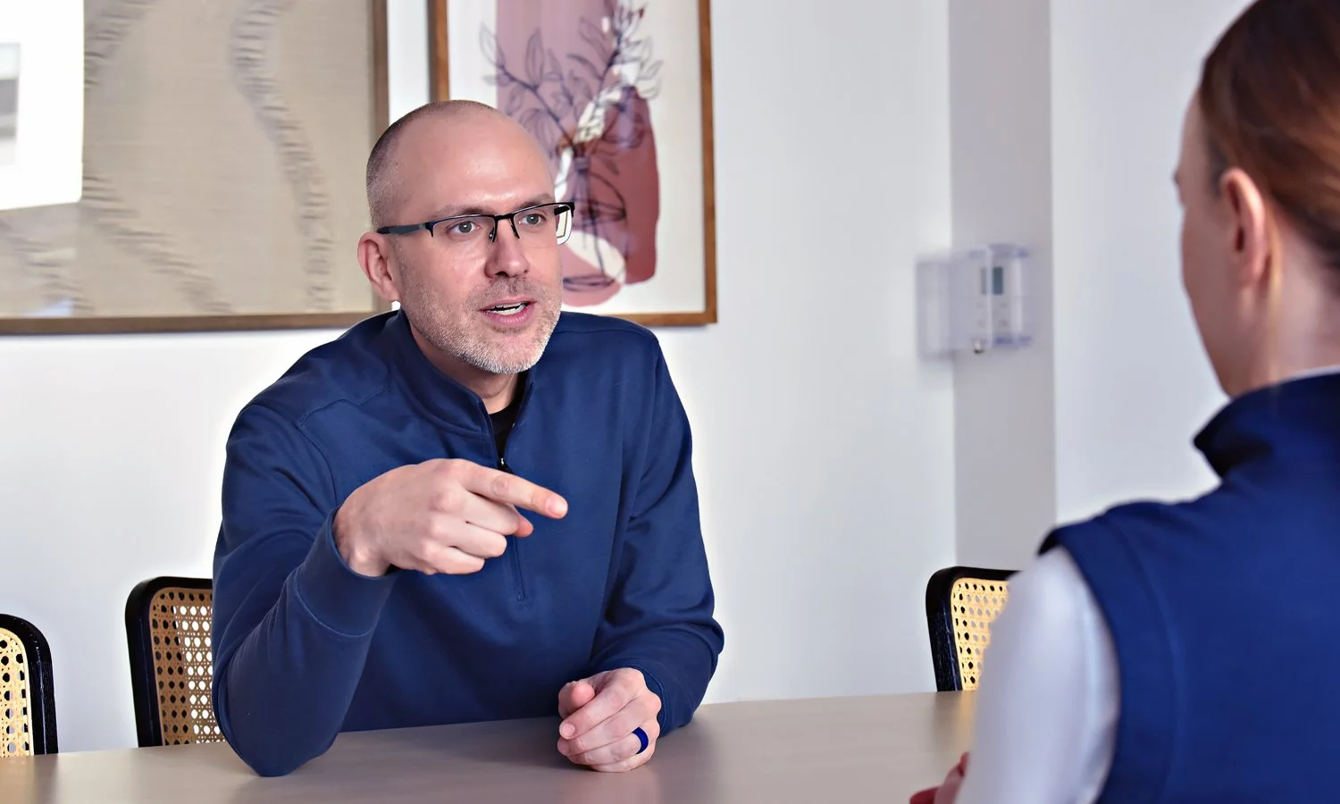 A man with glasses and a beard, wearing a blue jacket, points and talks to a woman with red hair, also in a blue jacket, in a modern office or conference room.