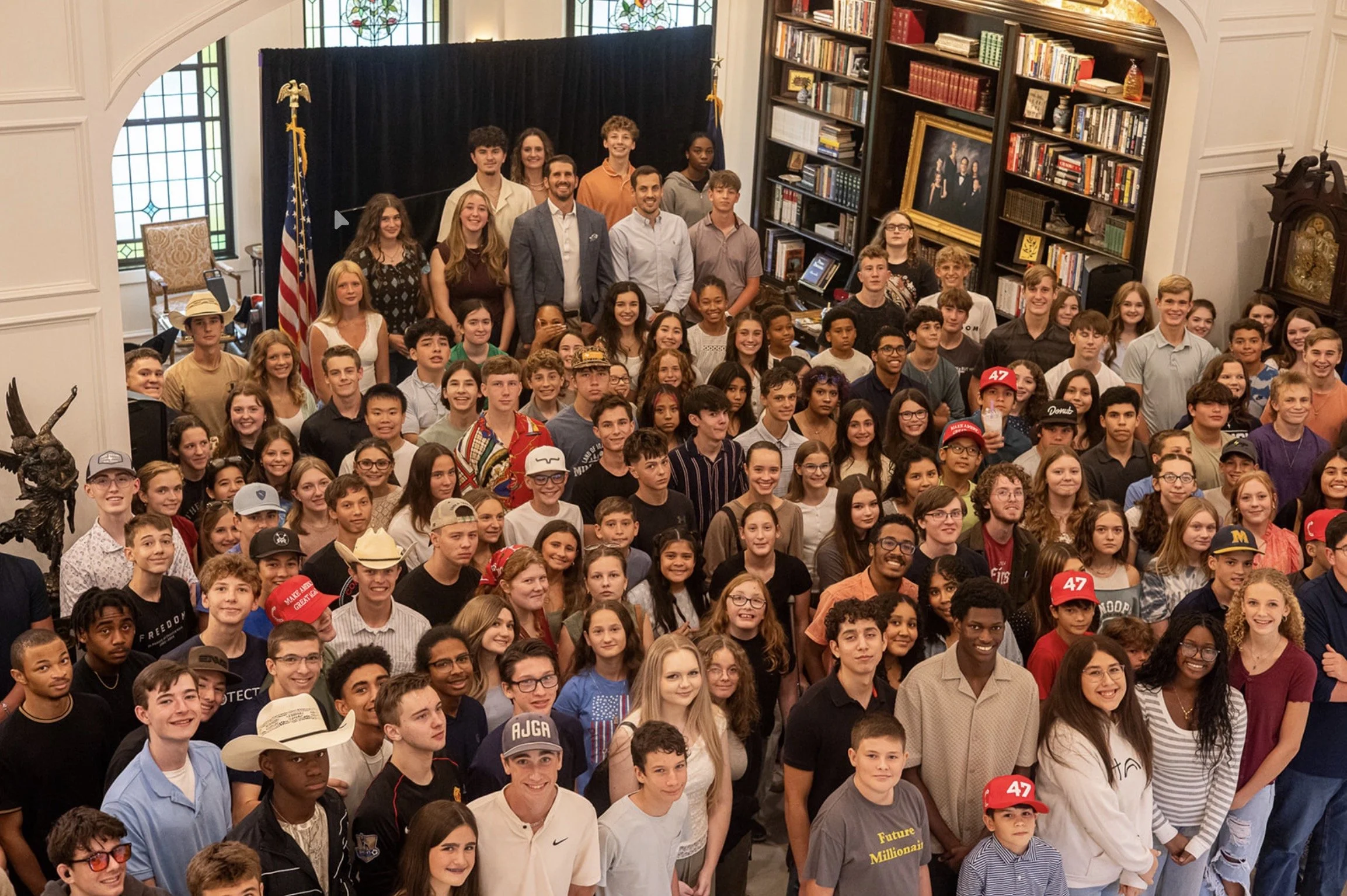 A large group of young people and adults gathered together indoors, smiling and posing for a group photo inside a room with bookshelves, stained glass windows, and flags.