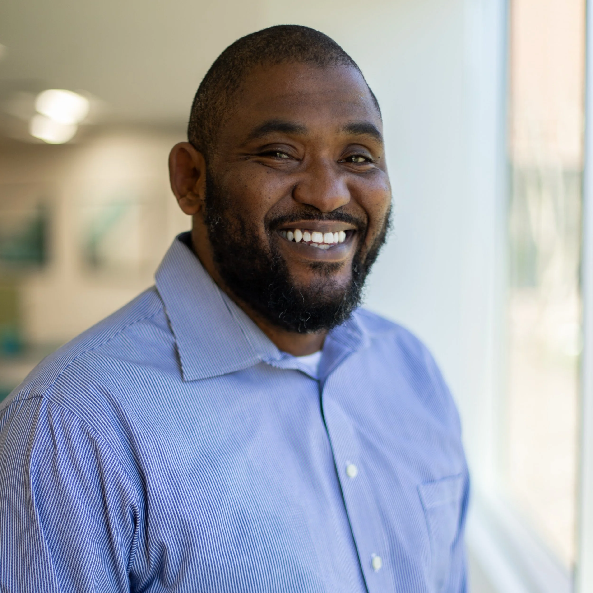 Close-up of a smiling African American man wearing a blue striped shirt in an indoor setting with blurred background, natural light.