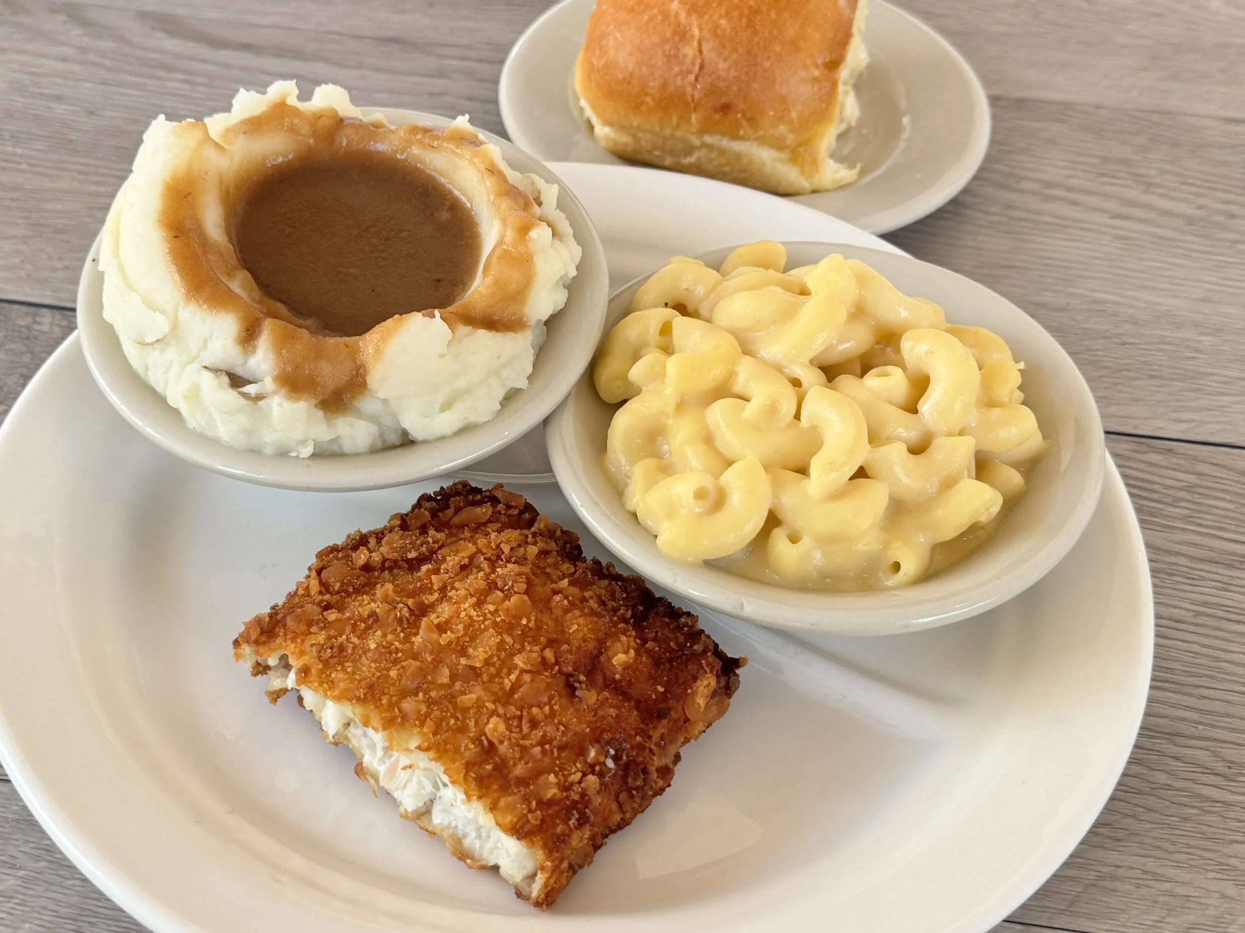 A dinner plate with mashed potatoes topped with gravy, macaroni and cheese, a baked bread roll, and a piece of fried chicken.