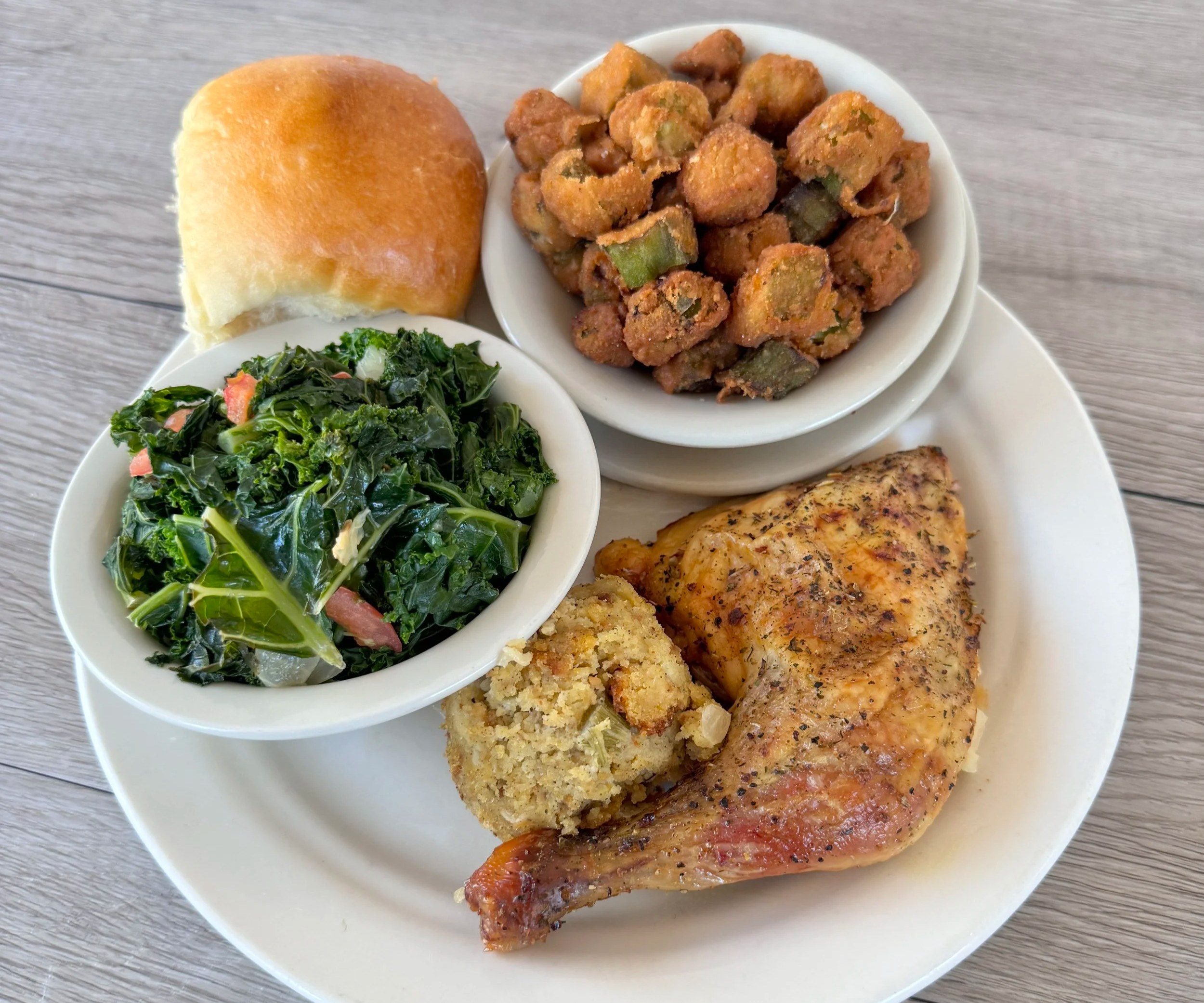 Plate with dinner items including a roll, a bowl of sautéed greens, baked chicken breast, cornbread, and a serving of fried okra.