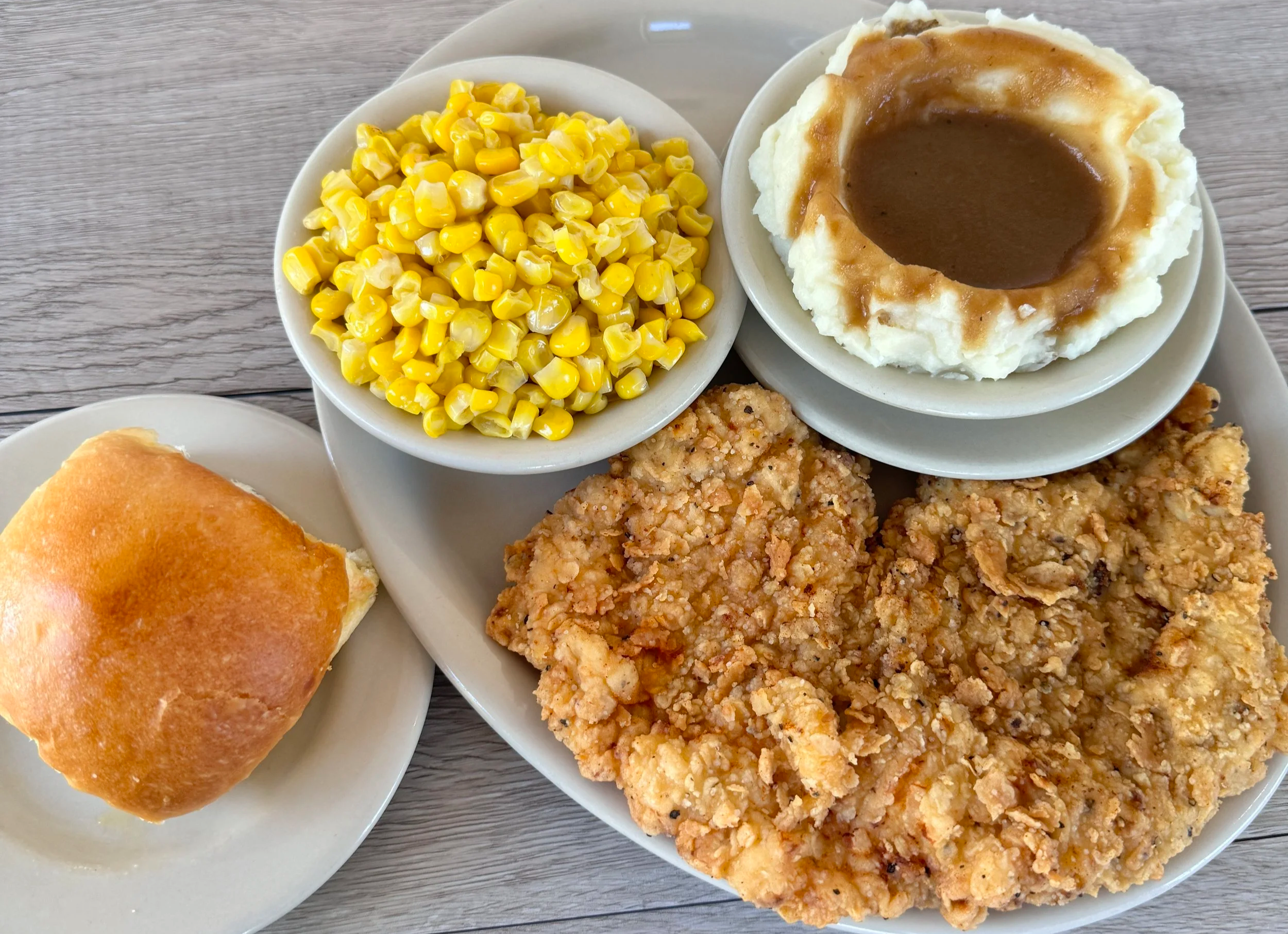 A plate with three pieces of fried chicken, a bowl of corn, a bread roll, and a bowl of mashed potatoes with brown gravy.