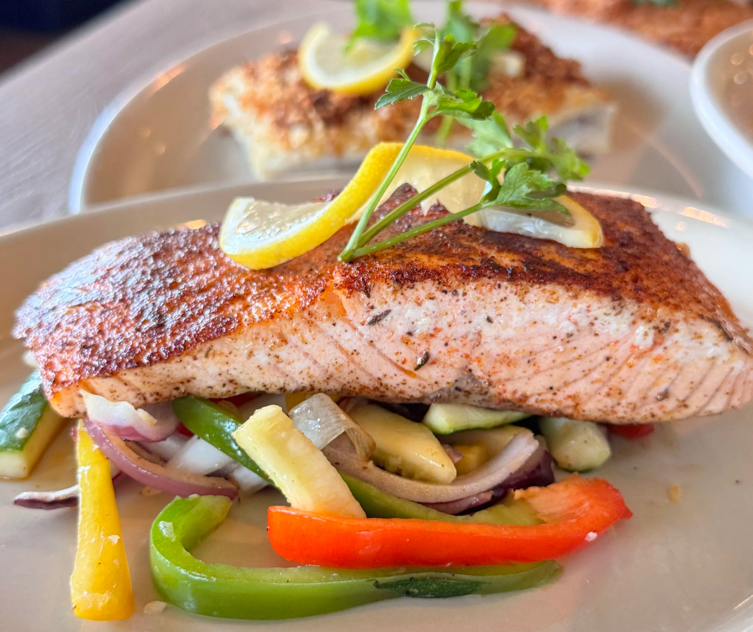 A plate of cooked salmon topped with lemon slices, fresh herbs, and a colorful vegetable medley including bell peppers, zucchini, and onions.