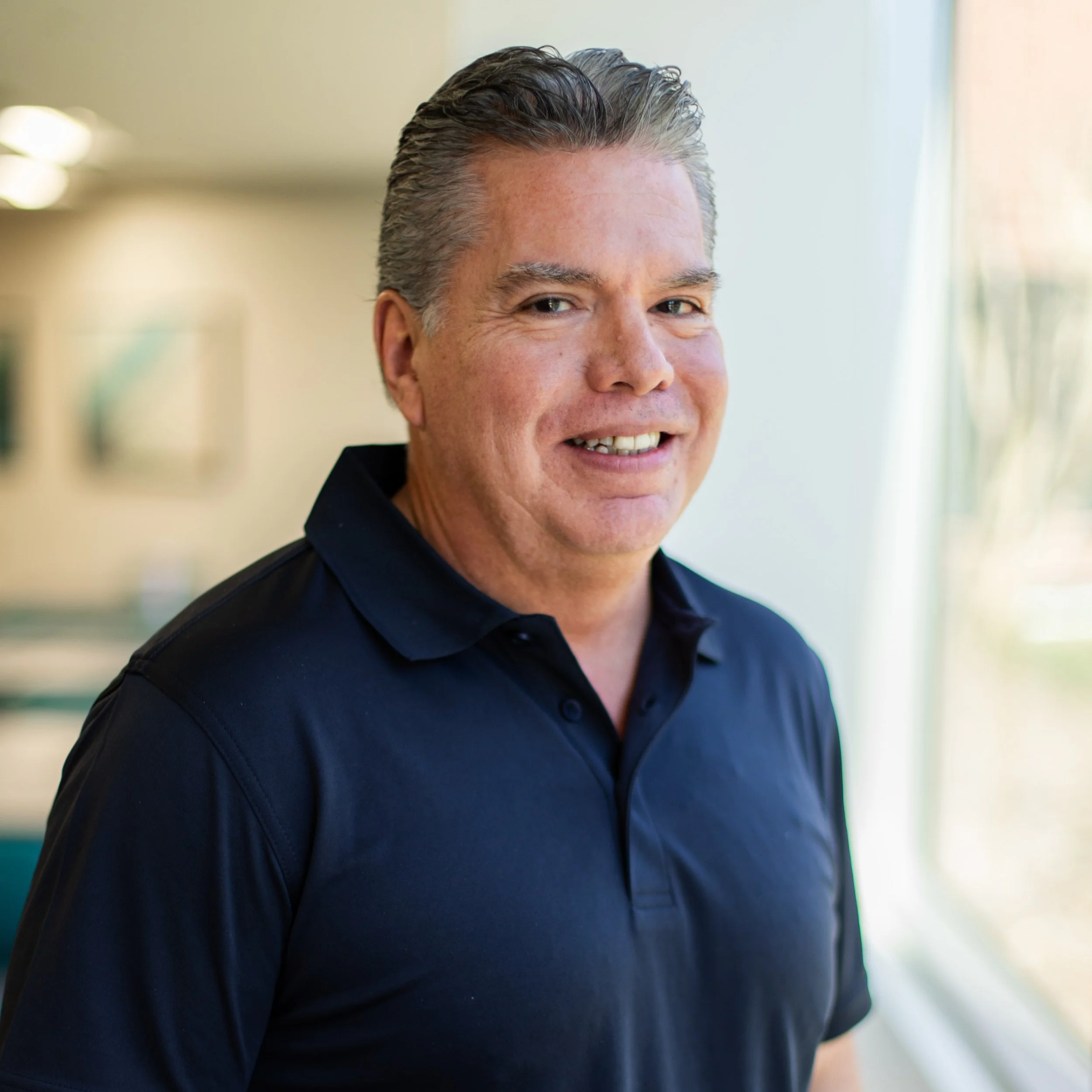 Close-up of a smiling middle-aged man with short, salt-and-pepper hair, wearing a black shirt, standing by a window with natural light.
