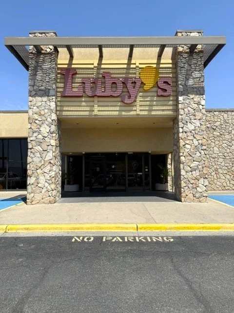 The entrance of a building with a sign that reads "Luby's" in pink and yellow letters, flanked by stone columns, with a "No Parking" sign painted on the pavement in front.
