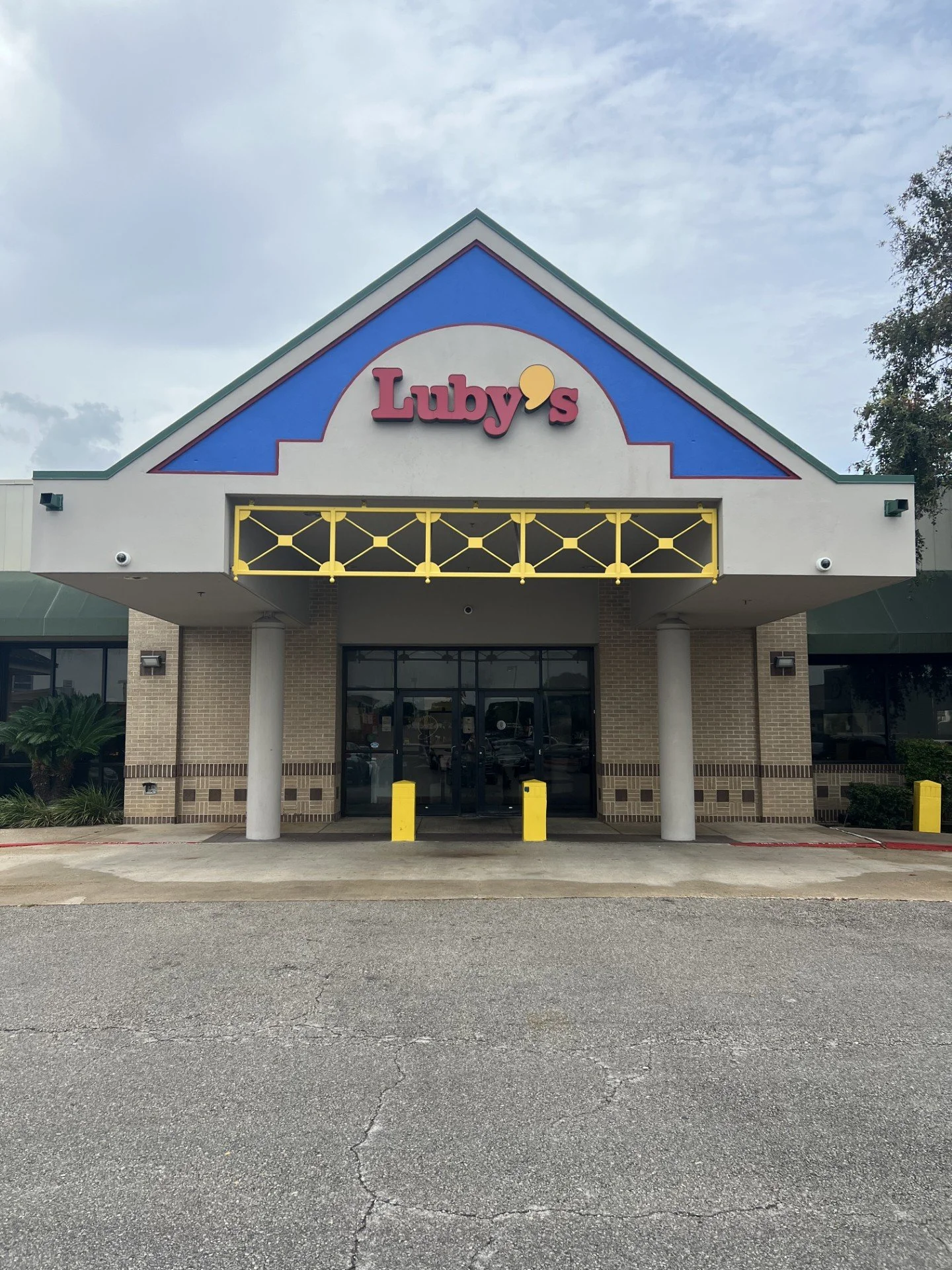 Front view of a shopping center entrance with a sign for Luby's, featuring a blue, white, and red color scheme, yellow decorative elements, two white columns, and parking lot in foreground with cracks.