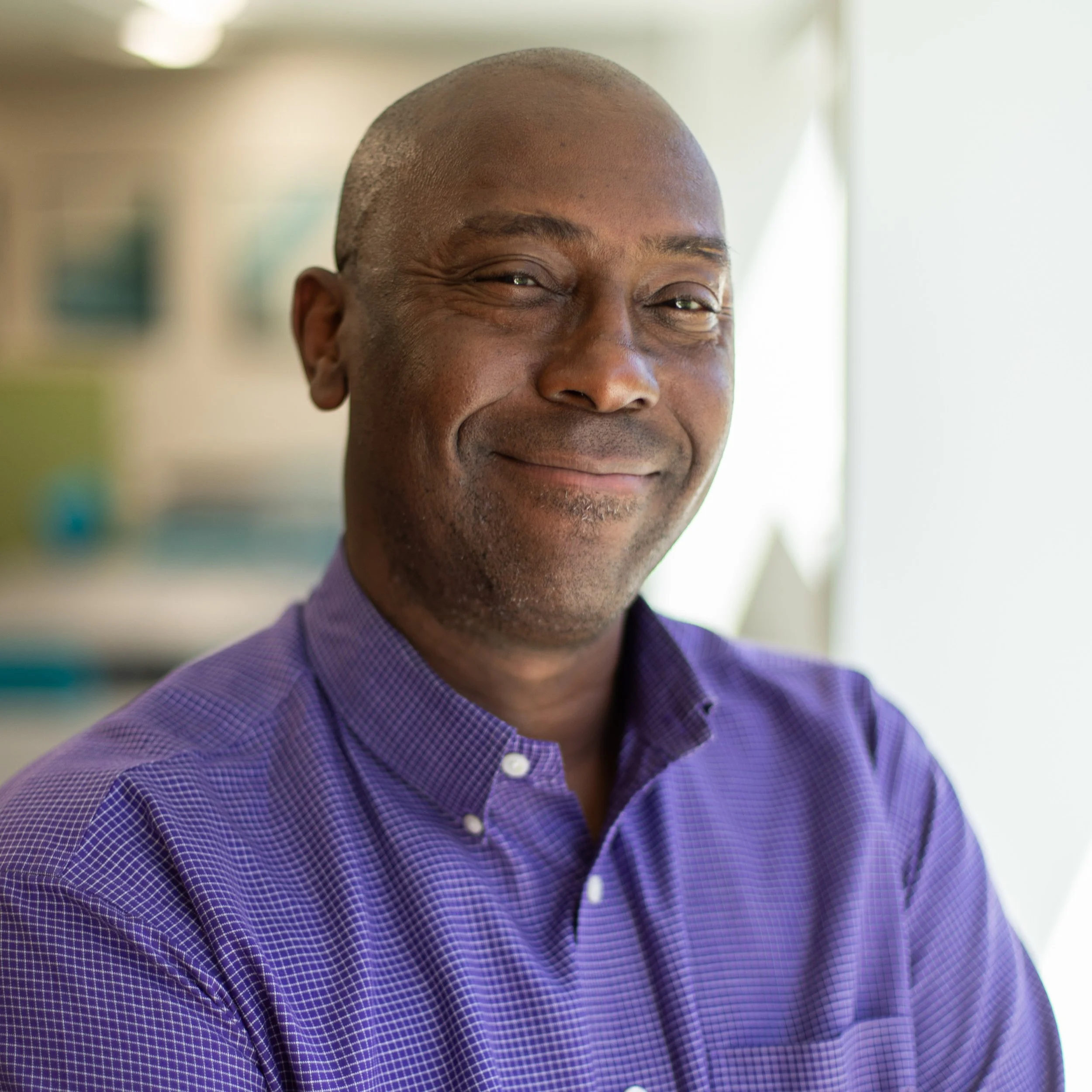 Close-up of a smiling African-American man with a shaved head wearing a purple checkered shirt in an indoor setting with a blurred background.