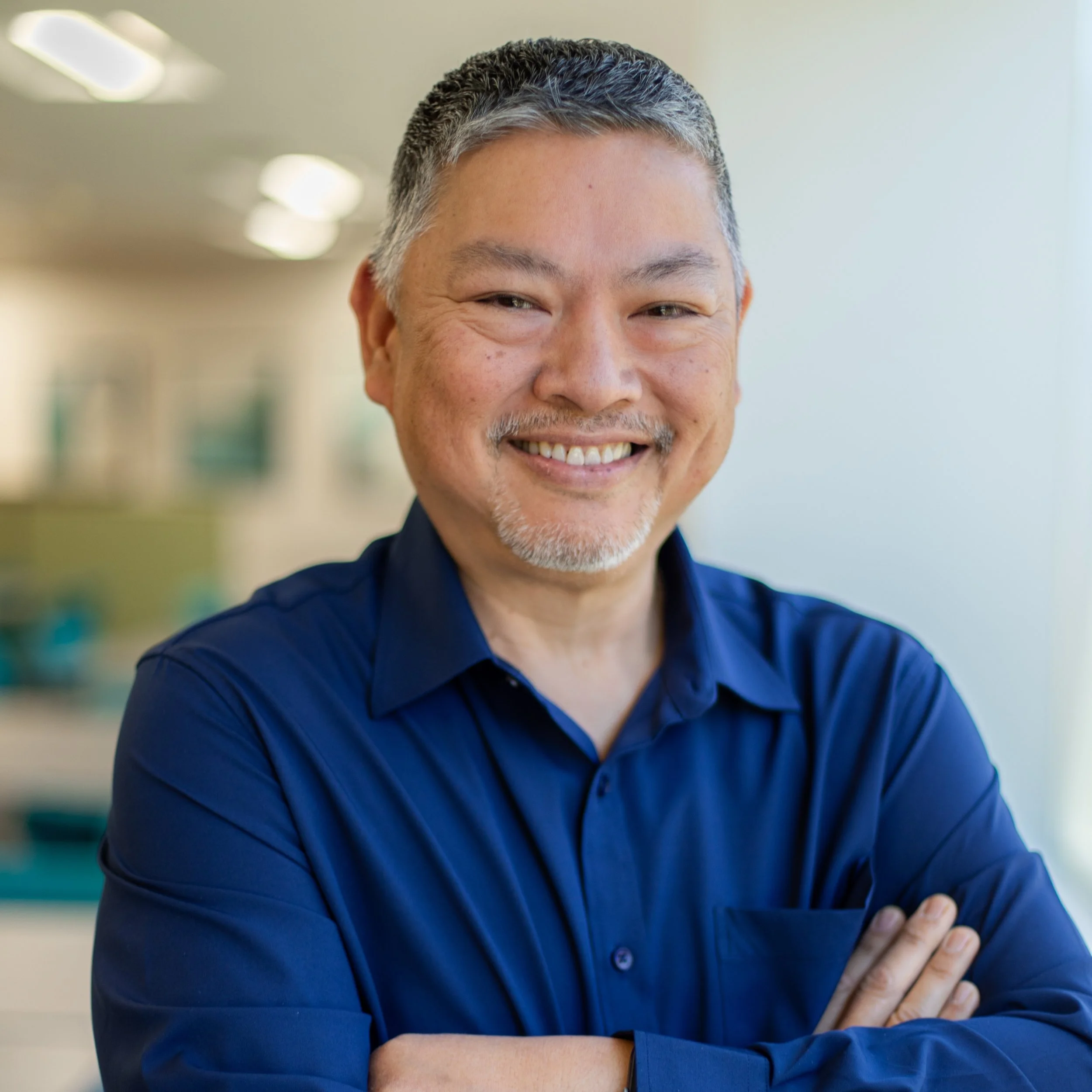 Close-up of a smiling middle-aged man with gray hair and a mustache, wearing a blue shirt, standing indoors with blurred background.