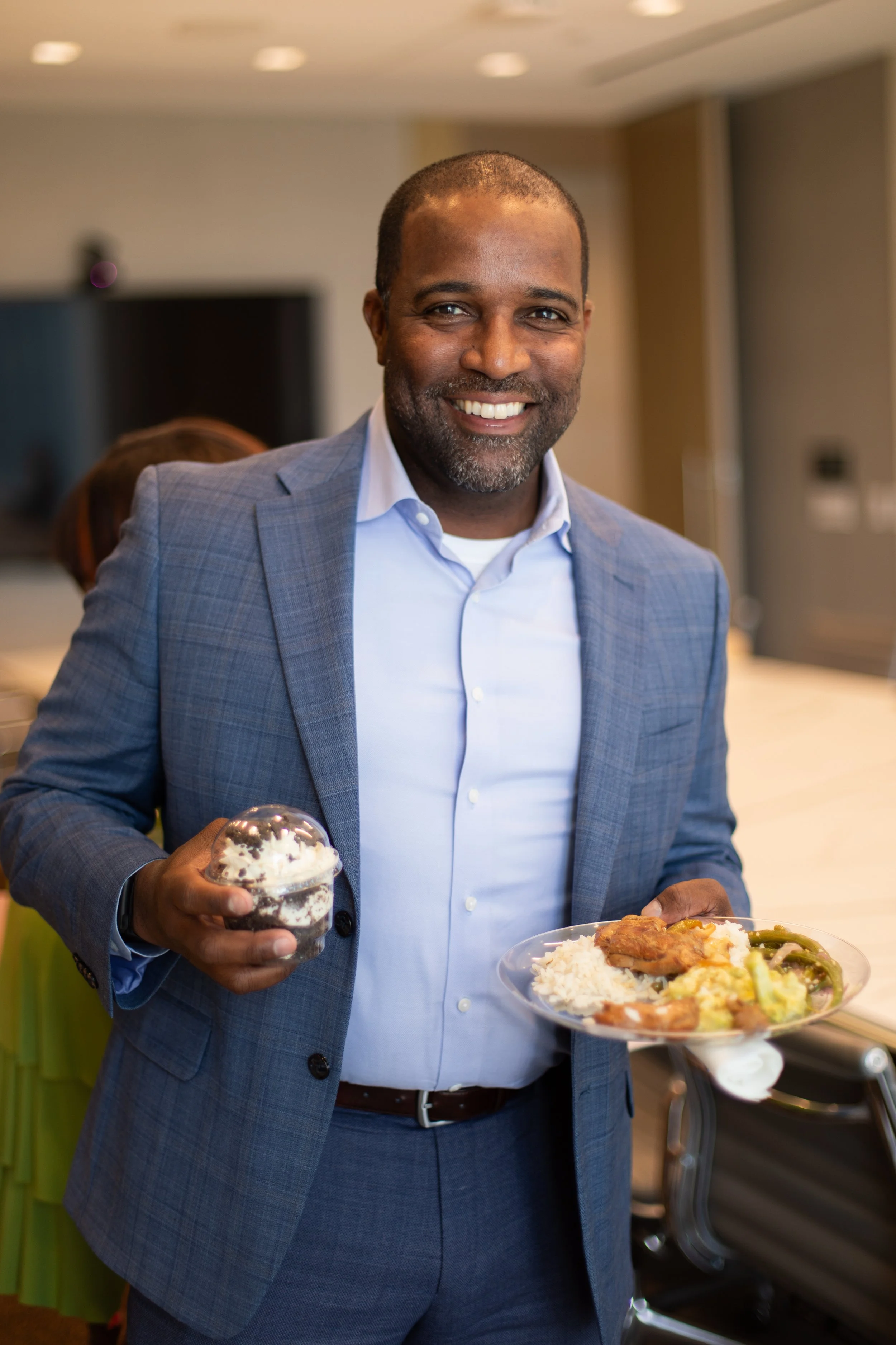 Man in a gray suit smiling at the camera while holding a dessert cup and a plate of food at a social gathering.