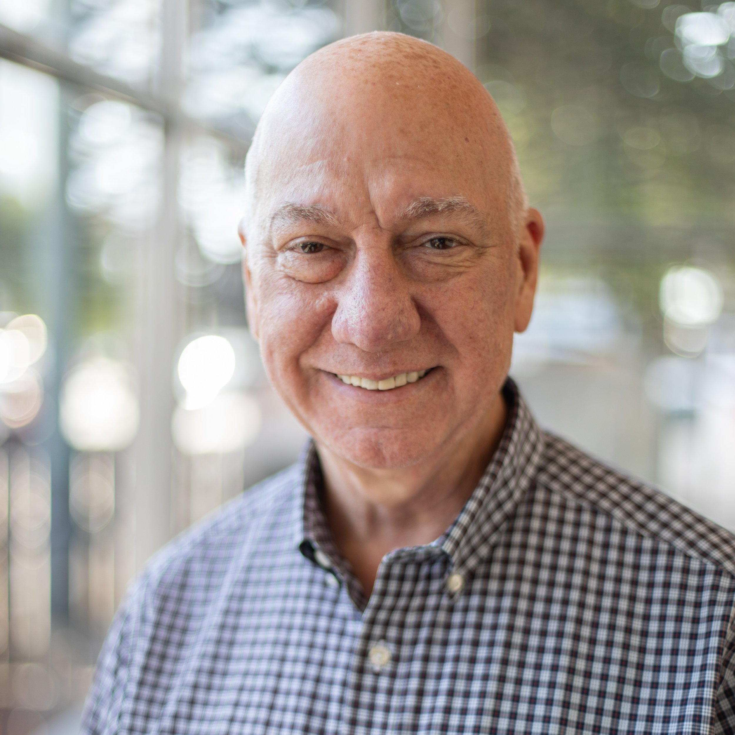 Close-up of a smiling elderly man with a bald head, wearing a checkered shirt, outdoors with blurred background.