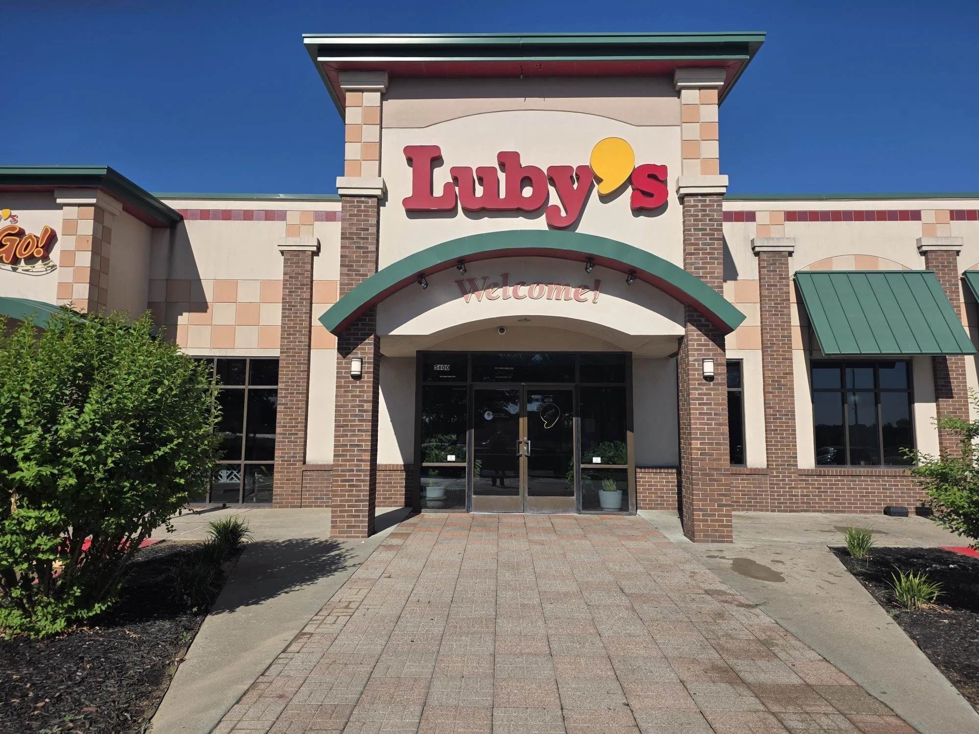 The storefront of a restaurant called Luby's with a welcoming sign, brick and stucco exterior, green roof accents, and a paved walkway leading to the entrance.