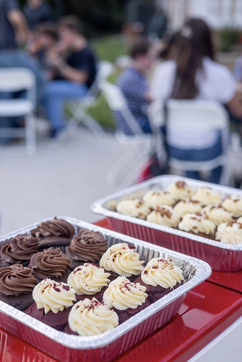 Tray of chocolate and vanilla cupcakes with frosting, outdoors with blurred group of people in the background.