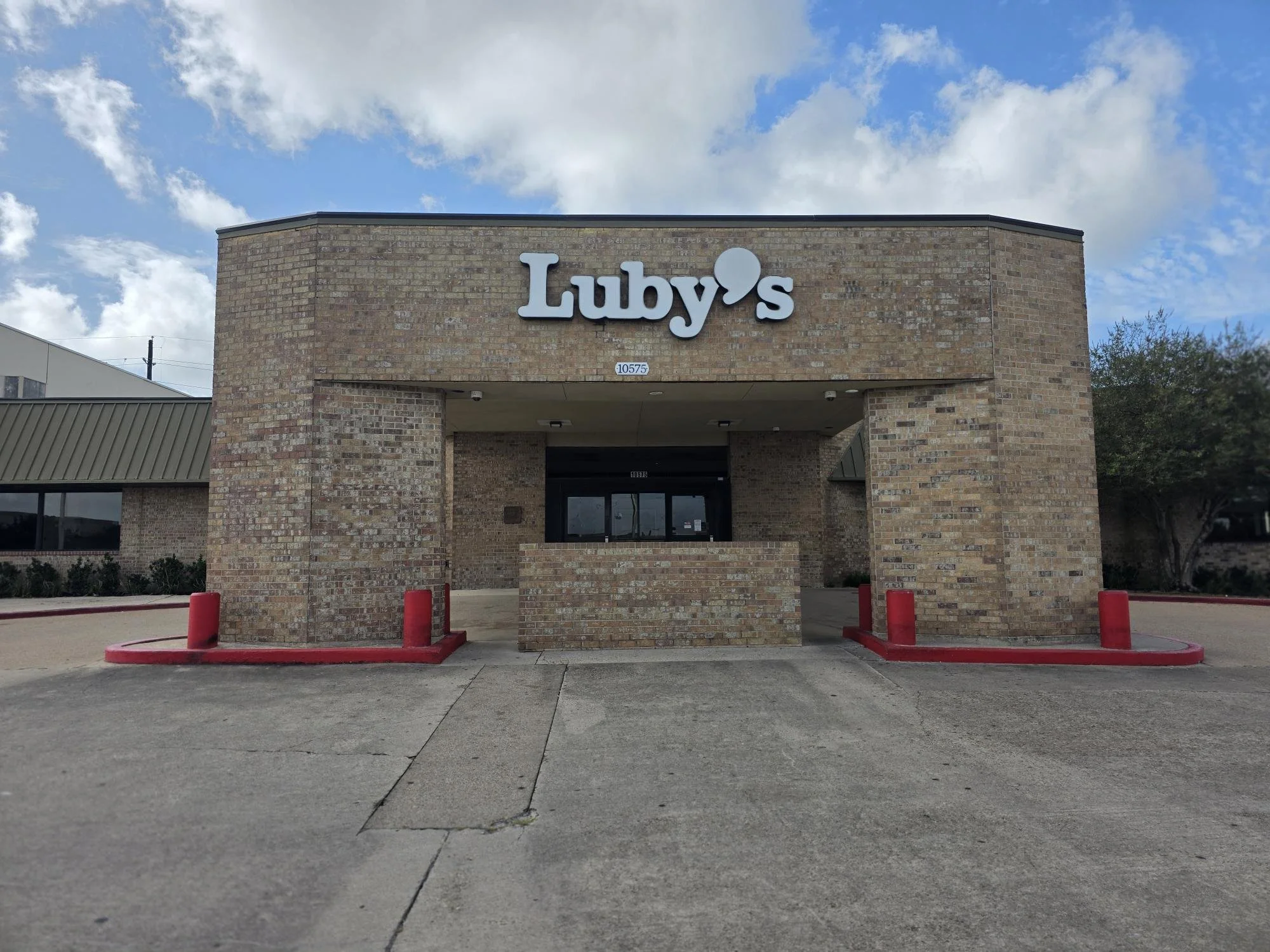 Exterior view of a brick building with a sign reading 'Luby's' above the entrance, under a partly cloudy sky. The parking area is in front with red-painted curbs and bollards.