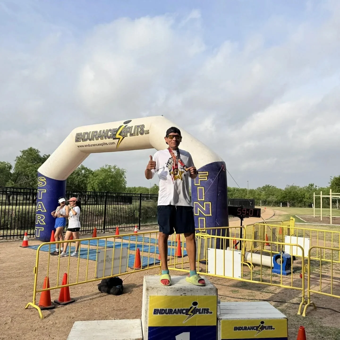 Male runner on the winners' podium at the end of a race, giving a thumbs-up. He is wearing sunglasses, a white T-shirt, blue shorts, and sandals. The background shows an inflatable arch with race branding, a digital clock, and a few spectators.