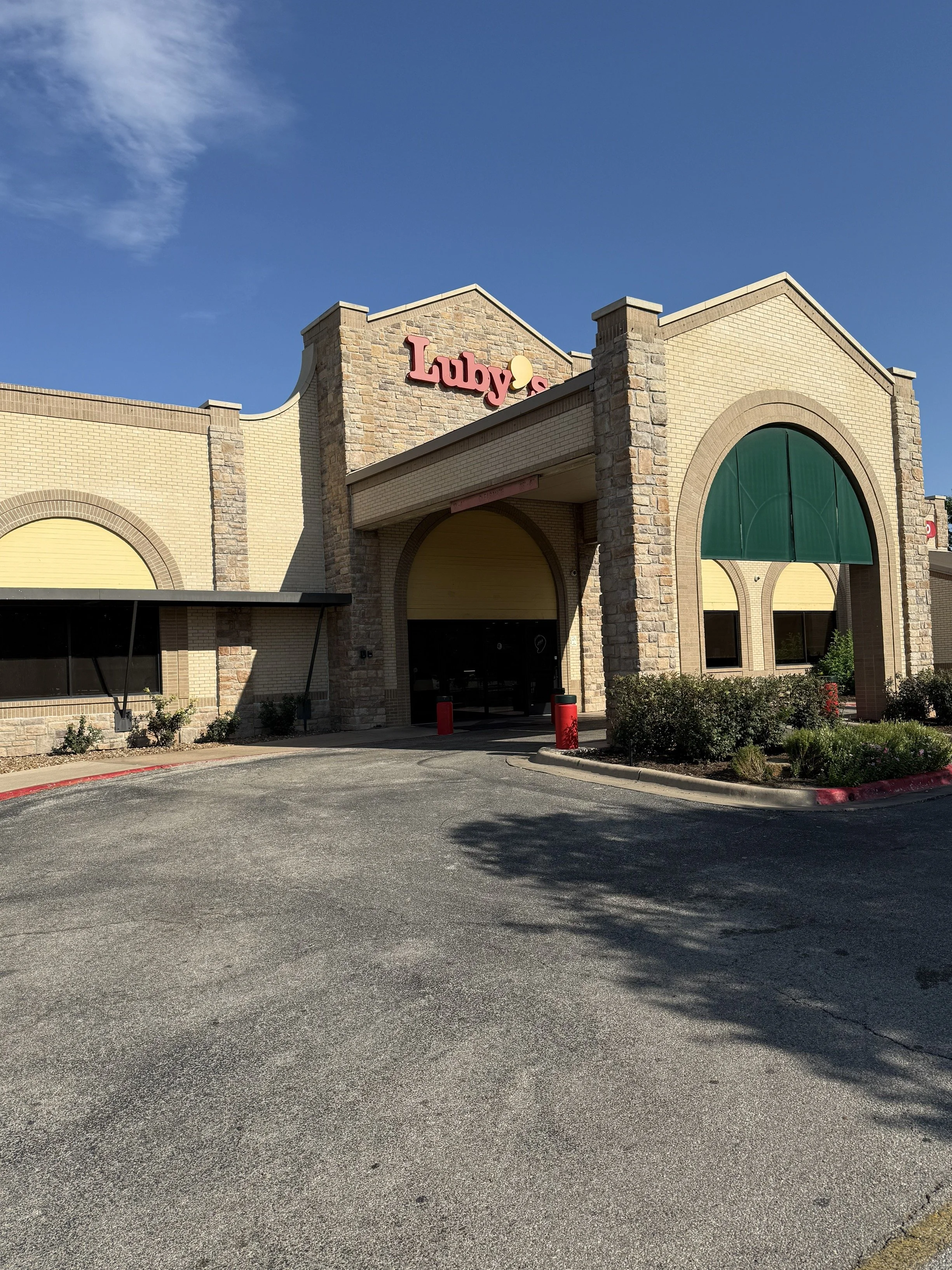 Exterior of a shopping center called Luby's with a brick facade and green awning on a sunny day.