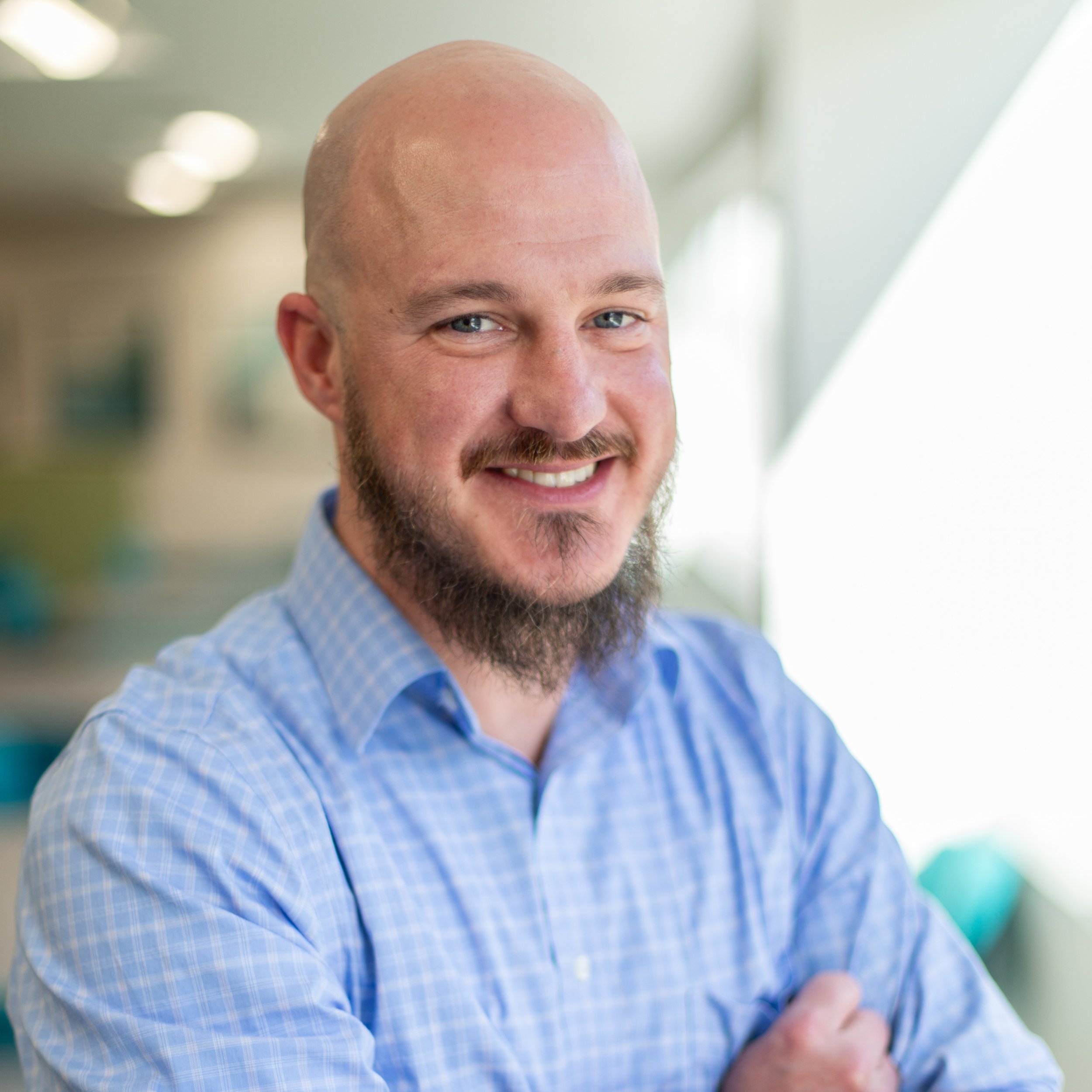 Close-up of a smiling man with a beard, blue eyes, and a bald head, wearing a blue checkered shirt, indoors with blurred background.