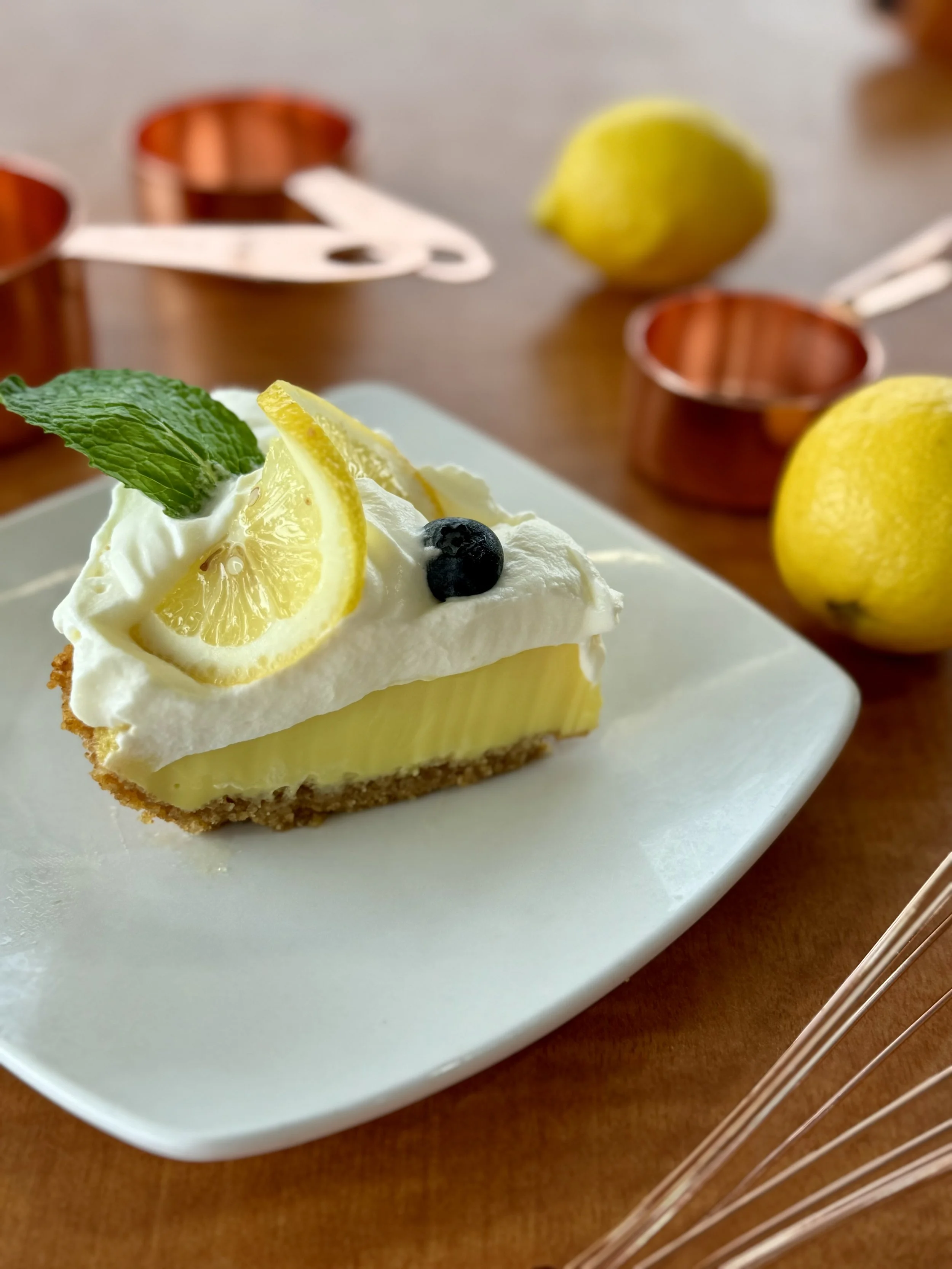 A slice of lemon dessert with a graham cracker crust, topped with whipped cream, lemon slices, a mint leaf, and blueberries, on a white plate with copper measuring cups and lemons in the background.
