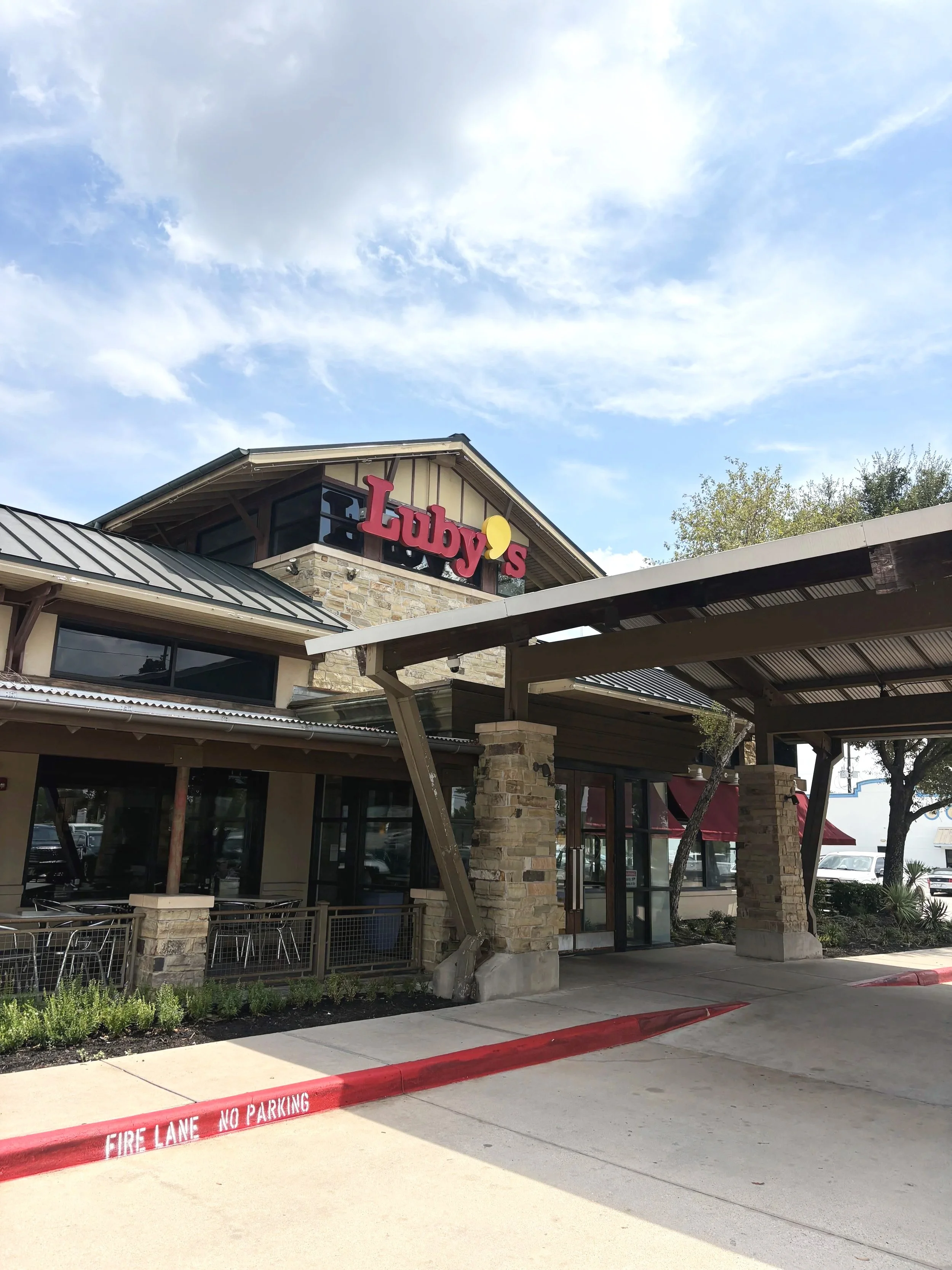 View of a building with a sign that reads 'Luby's', a patio area with tables and chairs, underneath a large metal awning, with a parking lot in front and a red curb with 'FIRE LANE NO PARKING' painted on it.