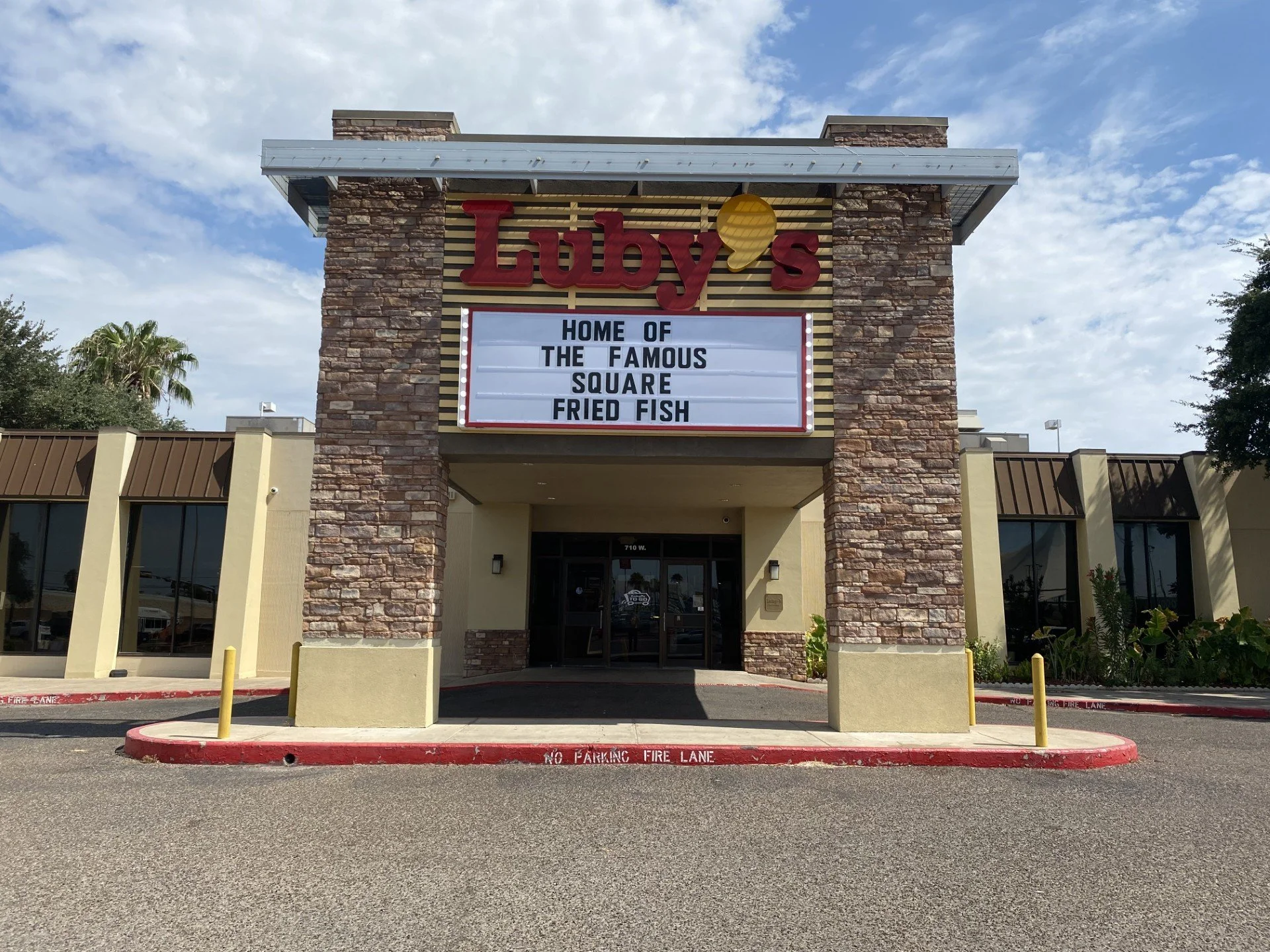 Front view of a brick building with a signboard stating "Luby's" in red letters, a yellow speech bubble, and a message about fried fish. The building is under a cloudy sky, with parking area in front and a covered walkway entrance.