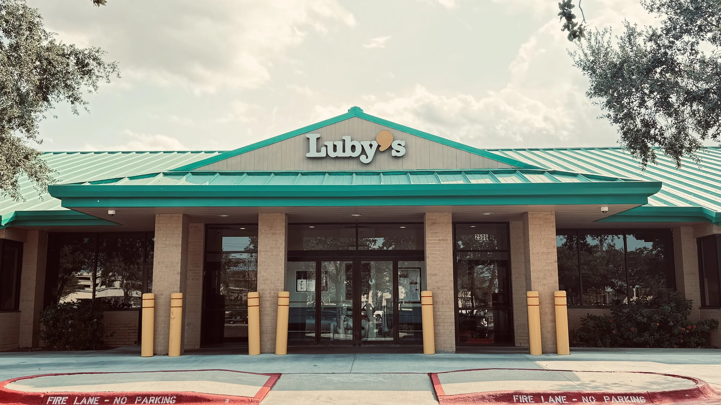 Luby's restaurant exterior with teal roof, brick walls, glass doors, and trees on sides.