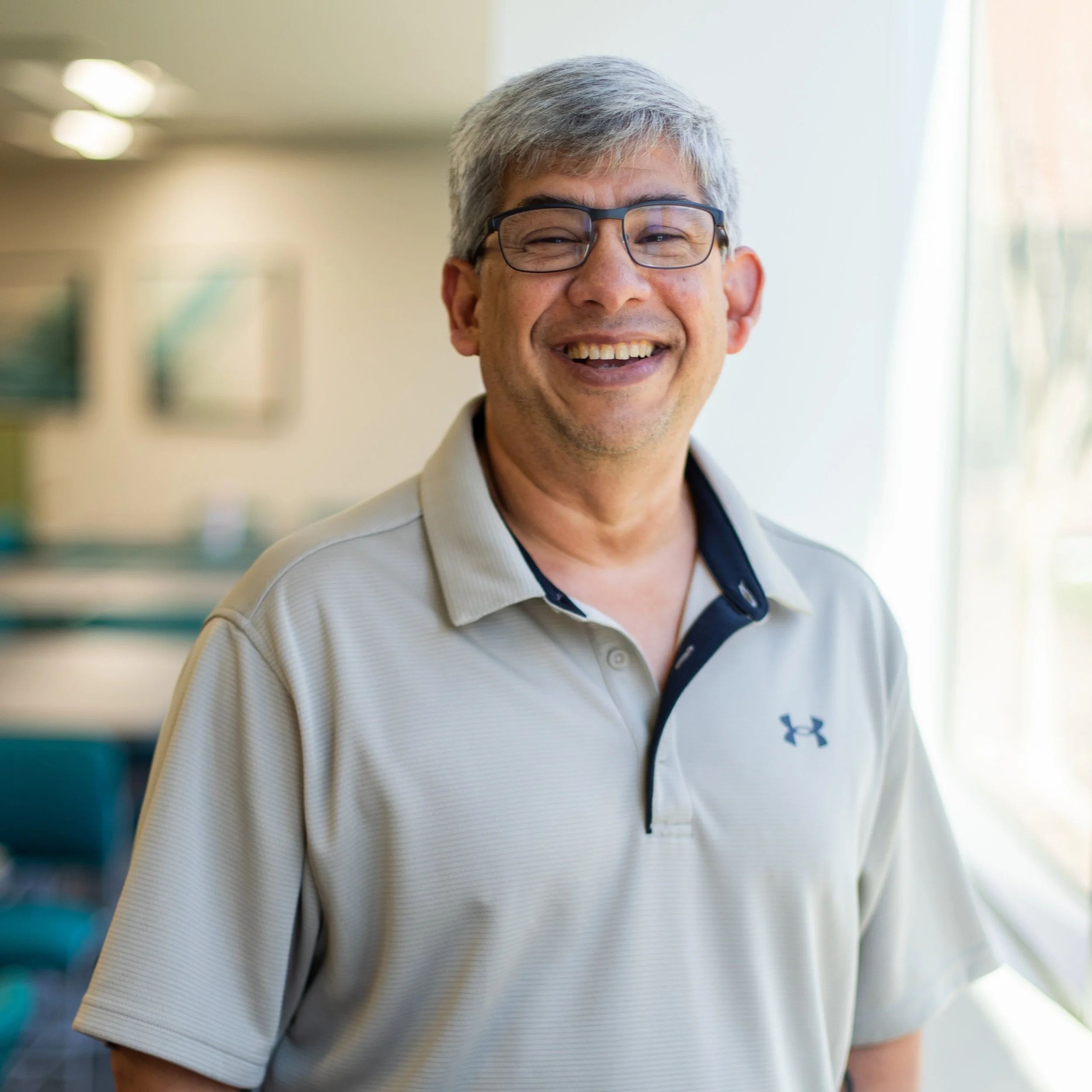 Close-up of a middle-aged man with gray hair and glasses, smiling warmly indoors, with a blurred background of bright and soft lighting.