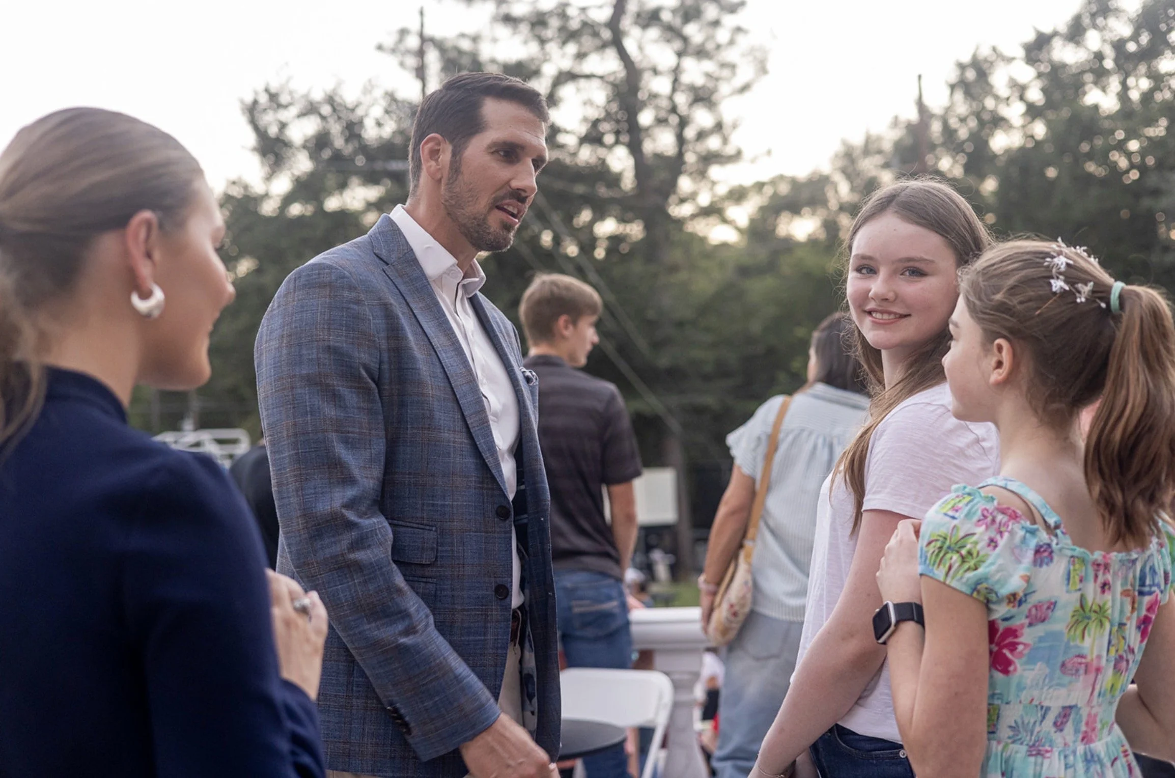 A man in a blue plaid suit speaking to two young girls with light brown hair at an outdoor event during the daytime, with other people and trees in the background.