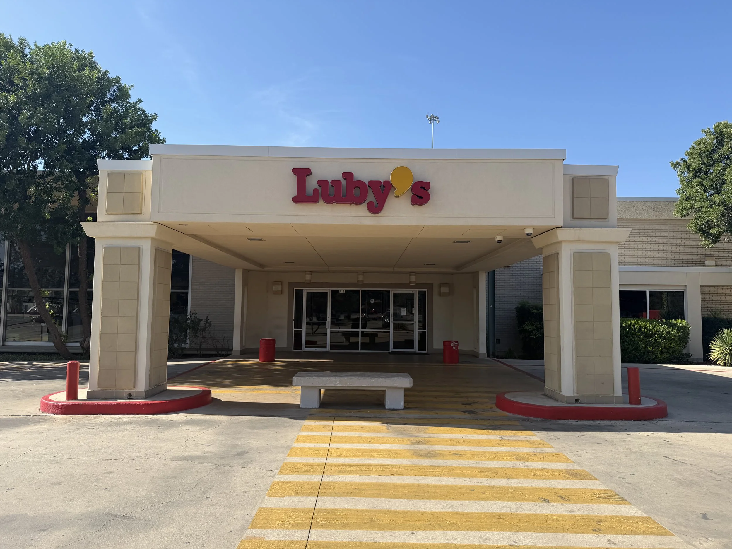 Entrance of a Luby's restaurant with a white facade, red sign, and yellow accent, featuring a covered walkway, surrounded by trees and a clear blue sky.