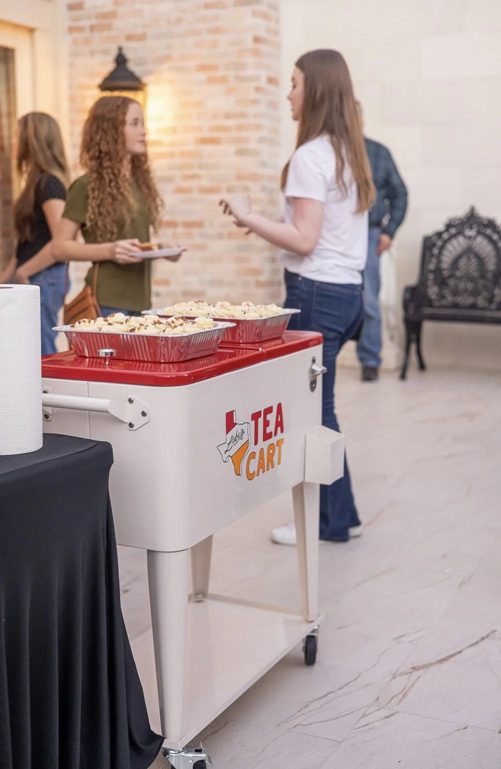 A white tea cart with red accents and a Texas-shaped logo labeled "Tea Cart" on a light-colored marble floor, with three large aluminum trays of food on top. In the background, several people are standing and talking near a brick wall with warm lighting.