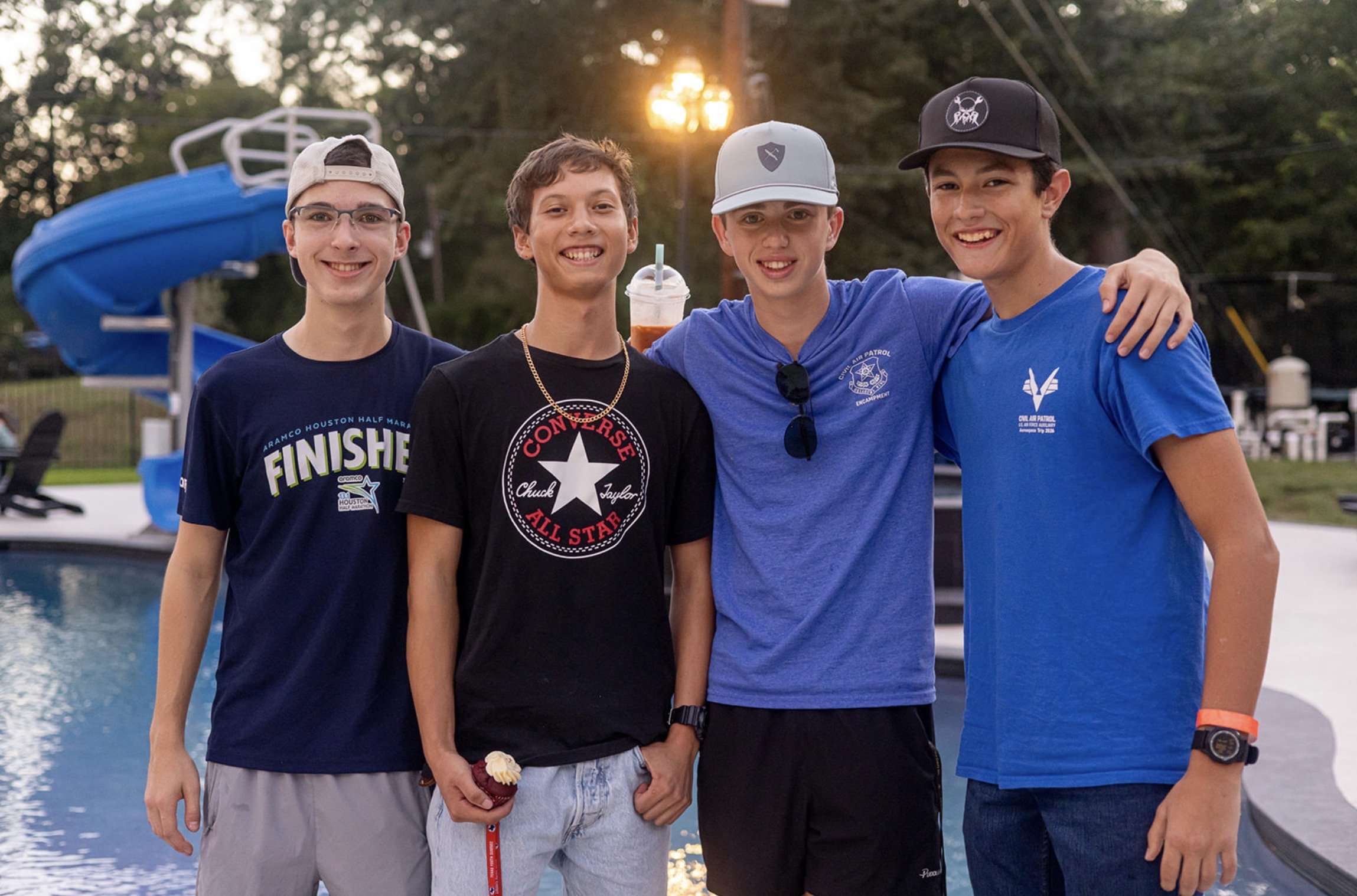 Four teenage boys standing side by side outdoors at sunset; they are smiling and have their arms around each other.