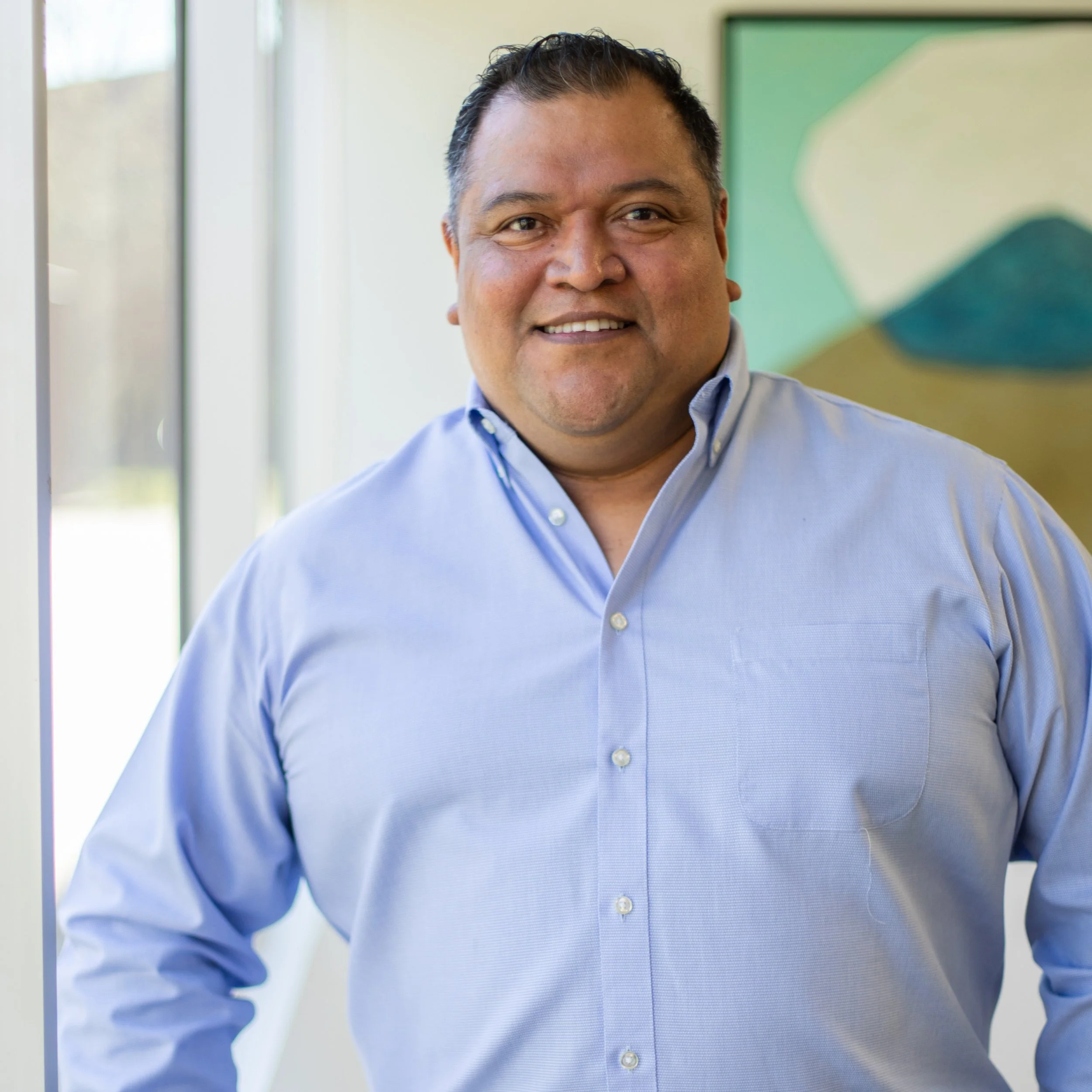 A smiling man in a light blue dress shirt standing indoors near a window and a green and blue abstract painting.