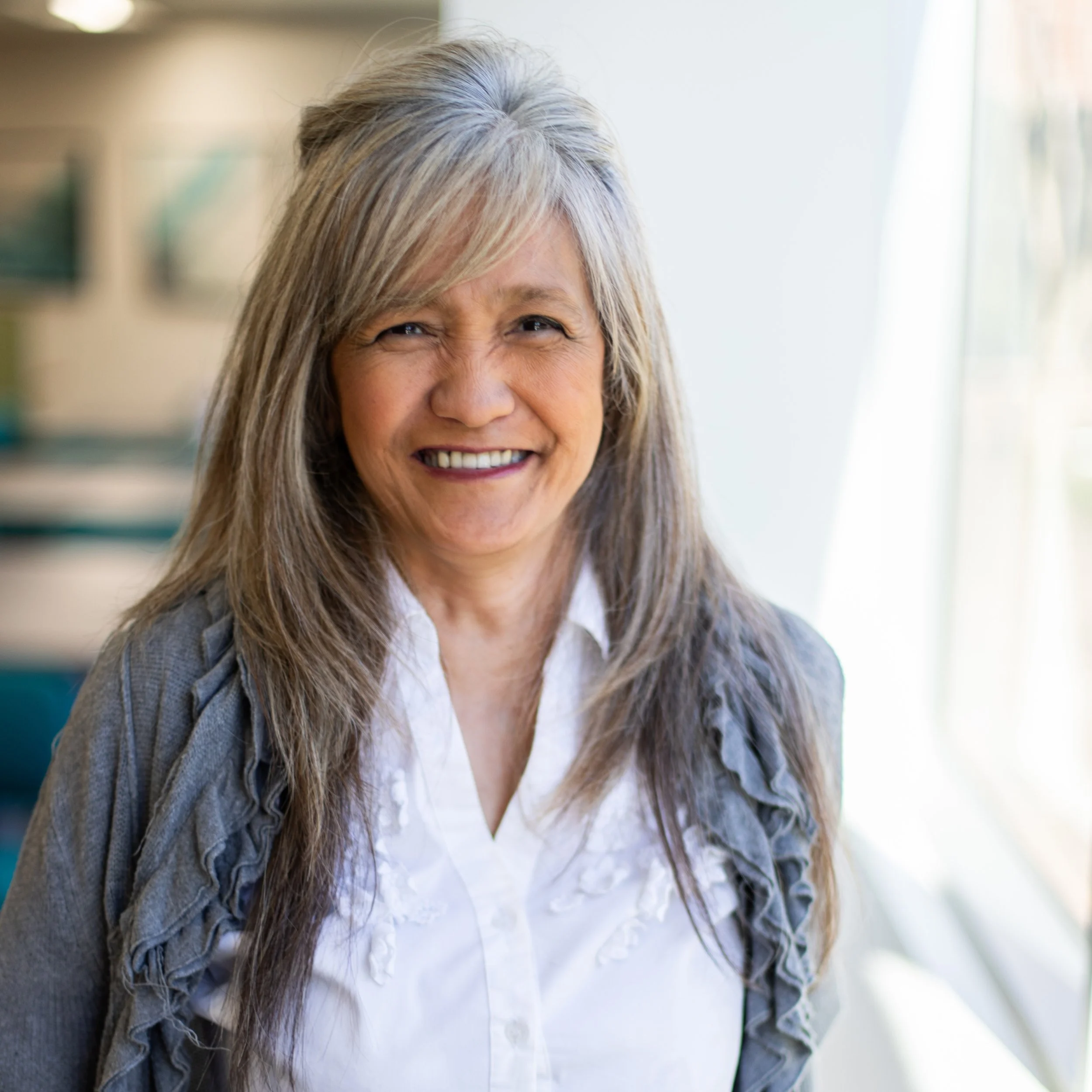 Close-up of an elderly woman smiling, with long gray hair and wearing a white shirt and a gray cardigan, standing next to a window with sunlight.