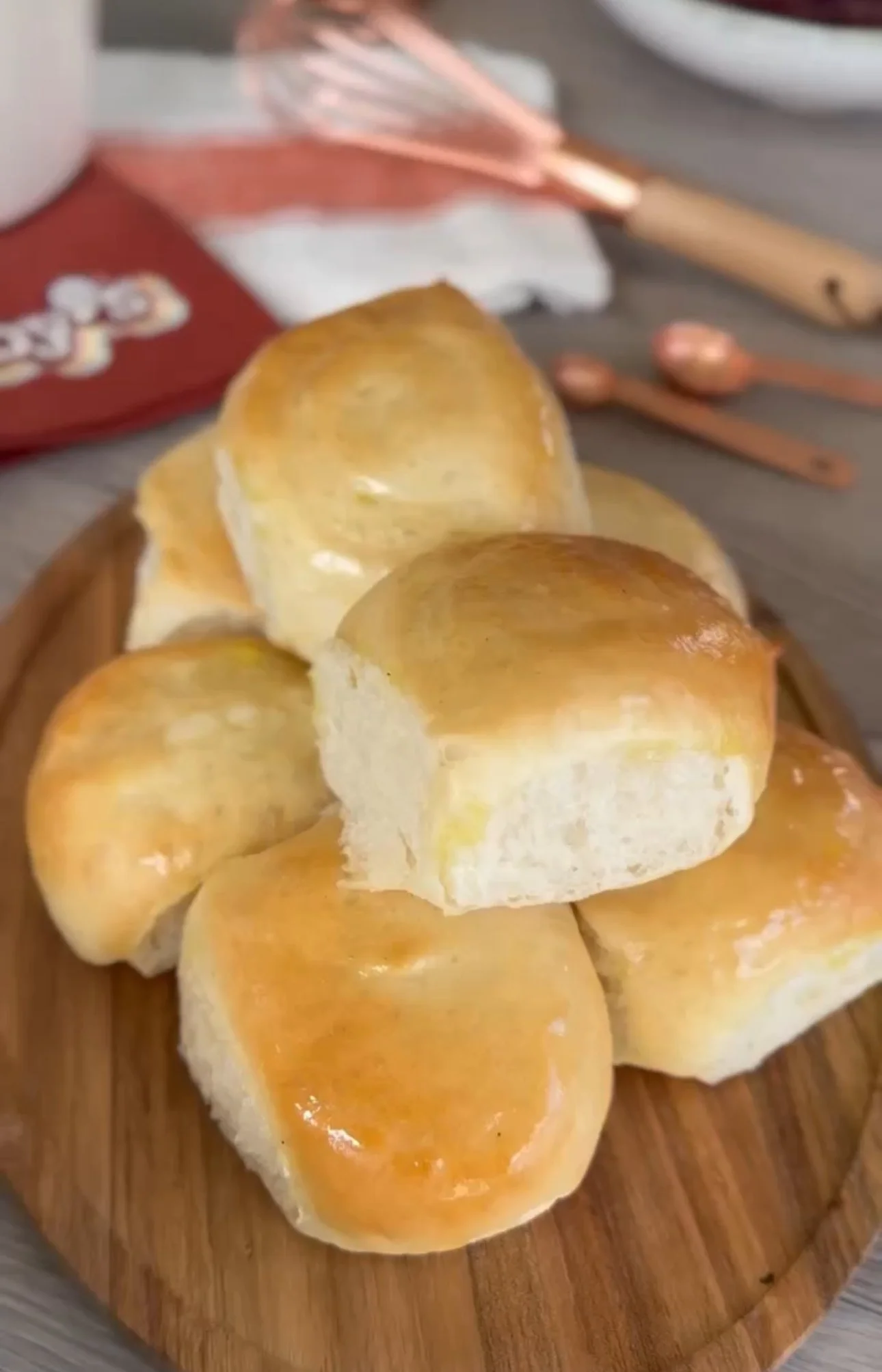 A wooden tray with freshly baked dinner rolls, some with a shiny, golden crust, on a wooden surface.