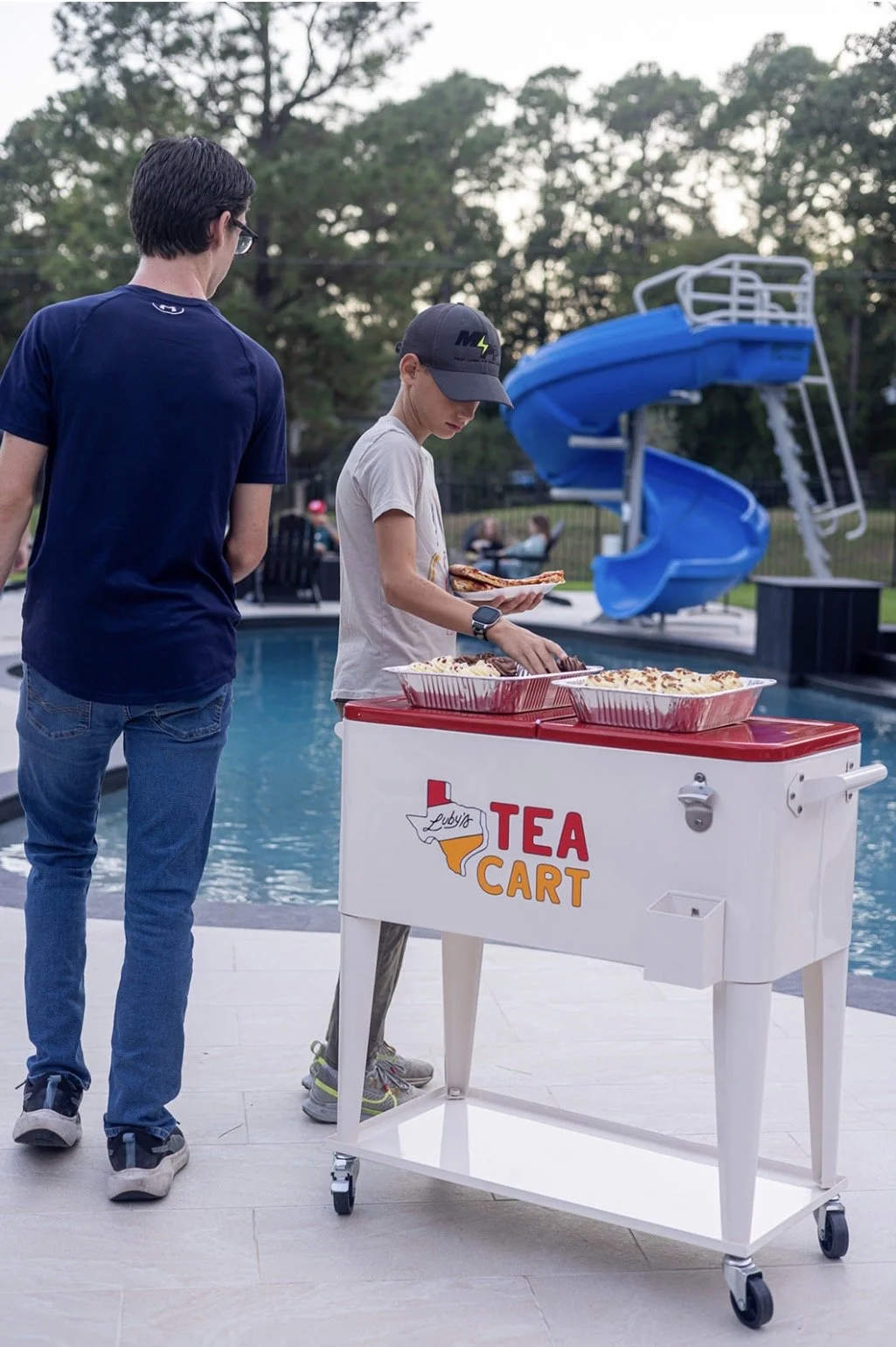 Two boys by a poolside with a white tea cart labeled "Tea Cart" serving pizza; one is selecting pizza while the other waits. In the background, there is a blue waterslide and people sitting on chairs.