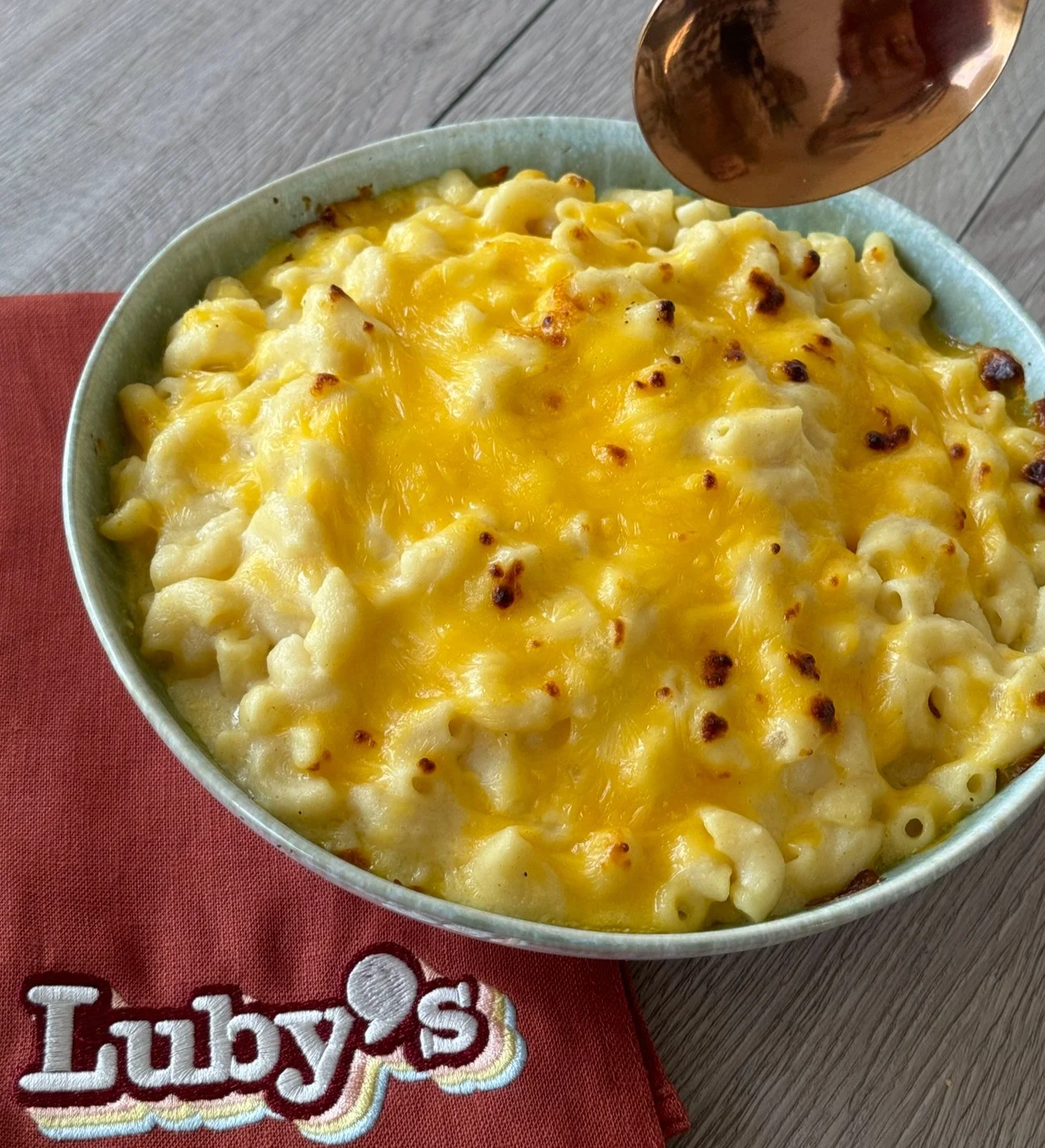 A bowl of baked macaroni and cheese with a golden melted cheese topping, placed on a red napkin with the 'Luby's' logo, on a wooden surface.