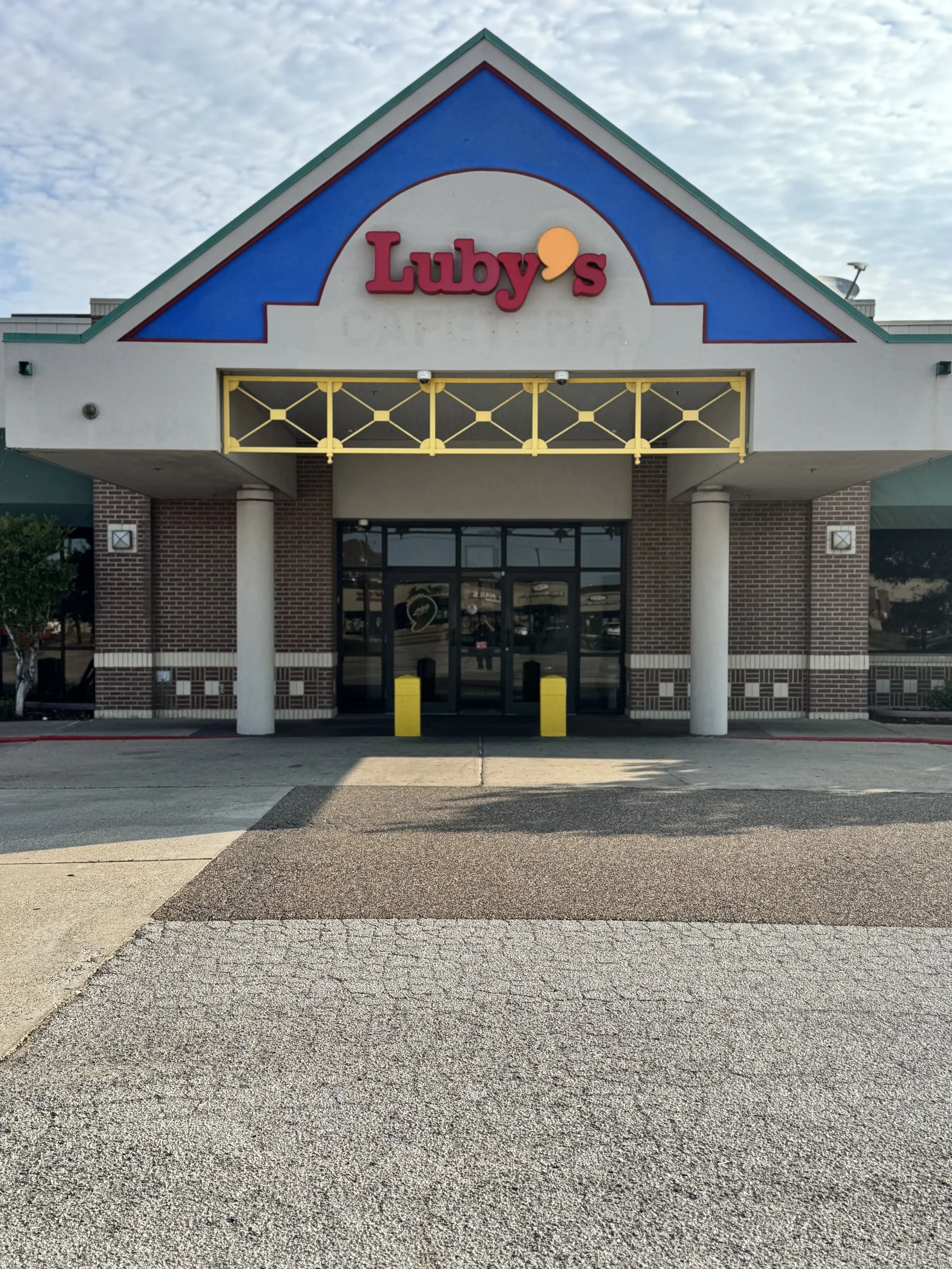 The exterior of Luby's restaurant with a brick facade, columns, and a blue, white, and green peaked roof. The Luby's sign, in red letters, is on the gable end of the roof.