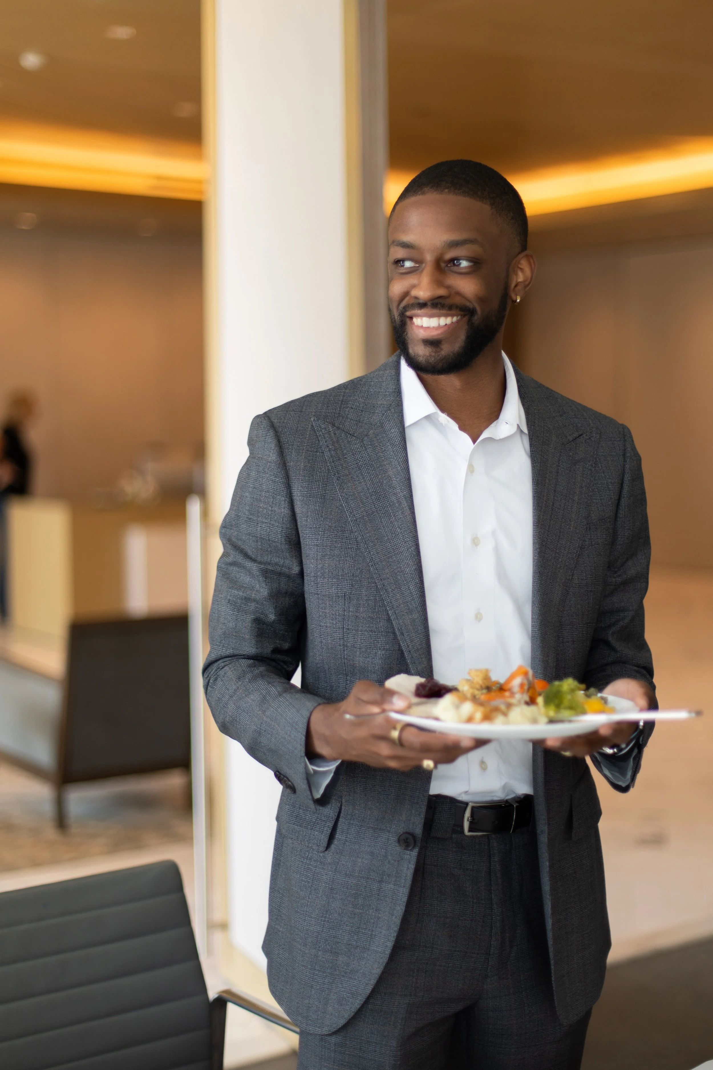 A smiling man in a gray business suit holding a plate of food in a modern indoor setting.