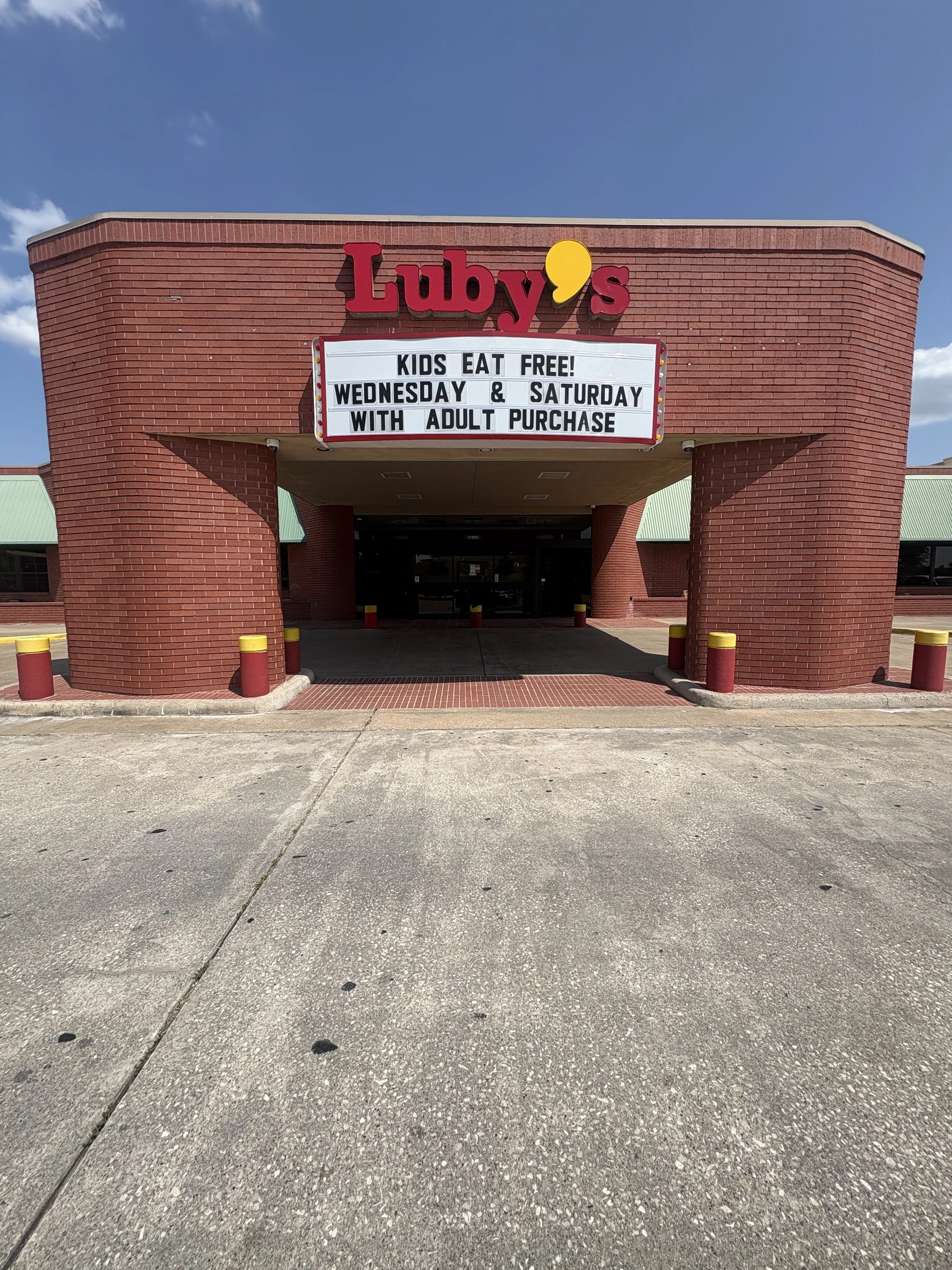 The entrance of a building with a red brick facade, a sign that says 'Luby's' in red and yellow letters, and a white message board displaying text about kids eat free on Wednesday and Saturday with adult purchase, under a blue sky.