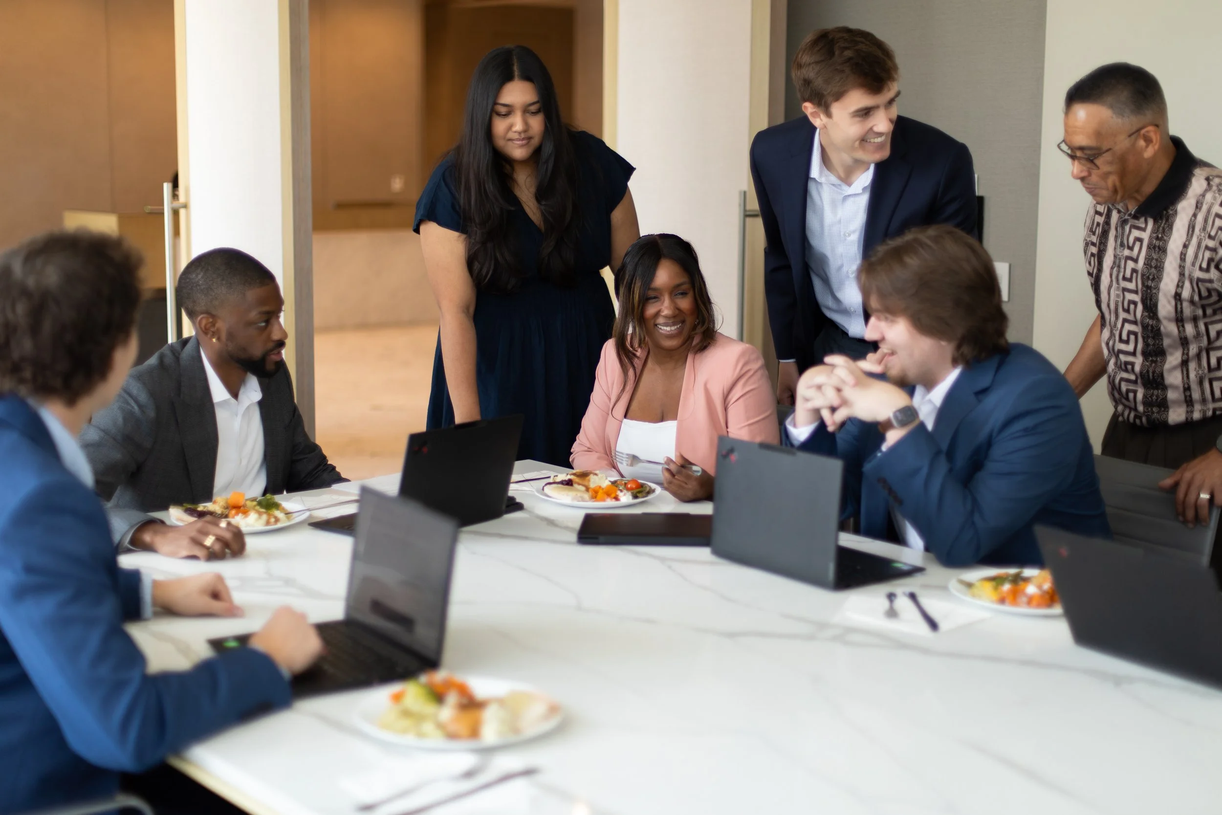 A diverse group of business professionals gathered around a conference table, engaged in a meeting with laptops and plates of food, some smiling and enjoying a discussion.