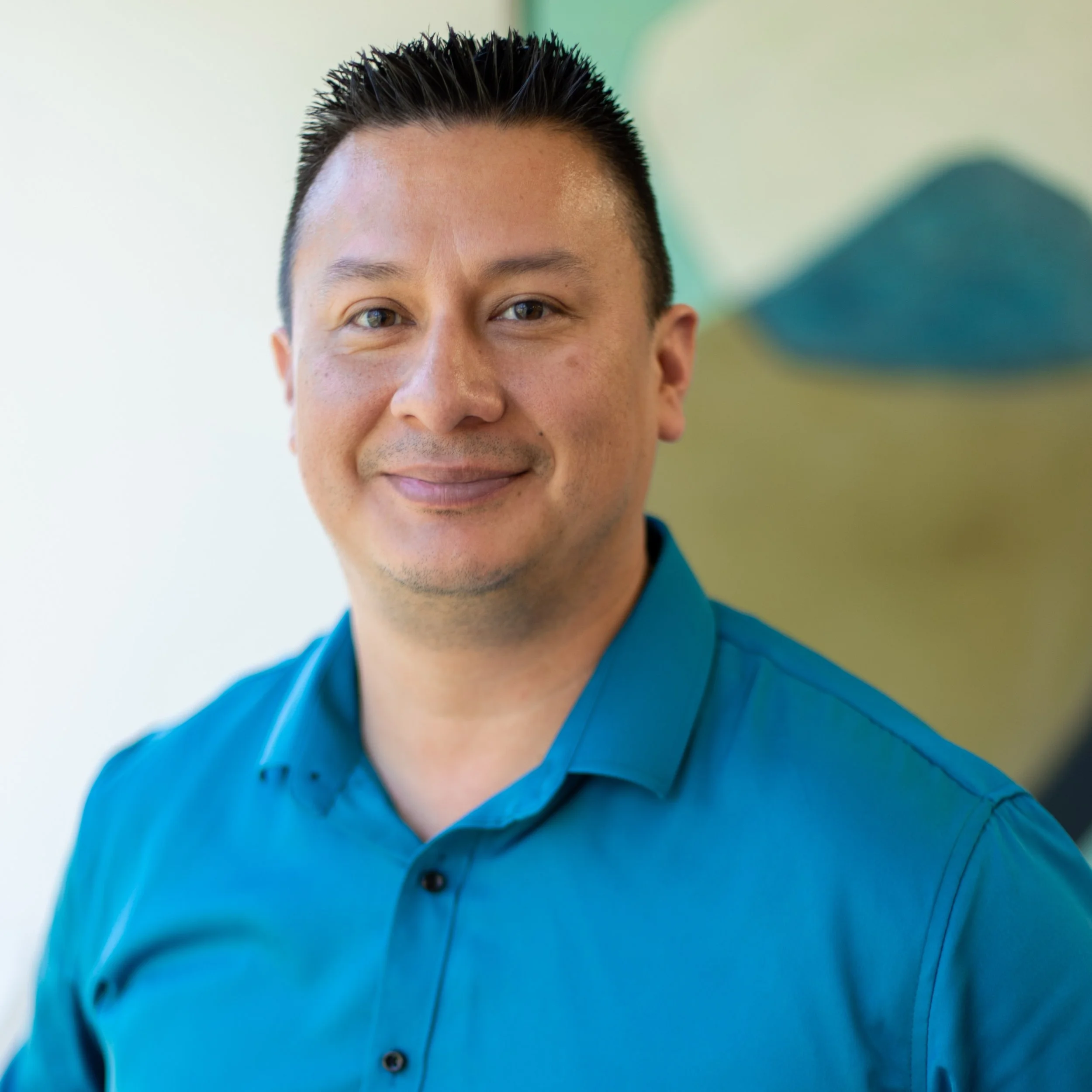 Close-up of a smiling man in a blue shirt against a blurred green and blue background.