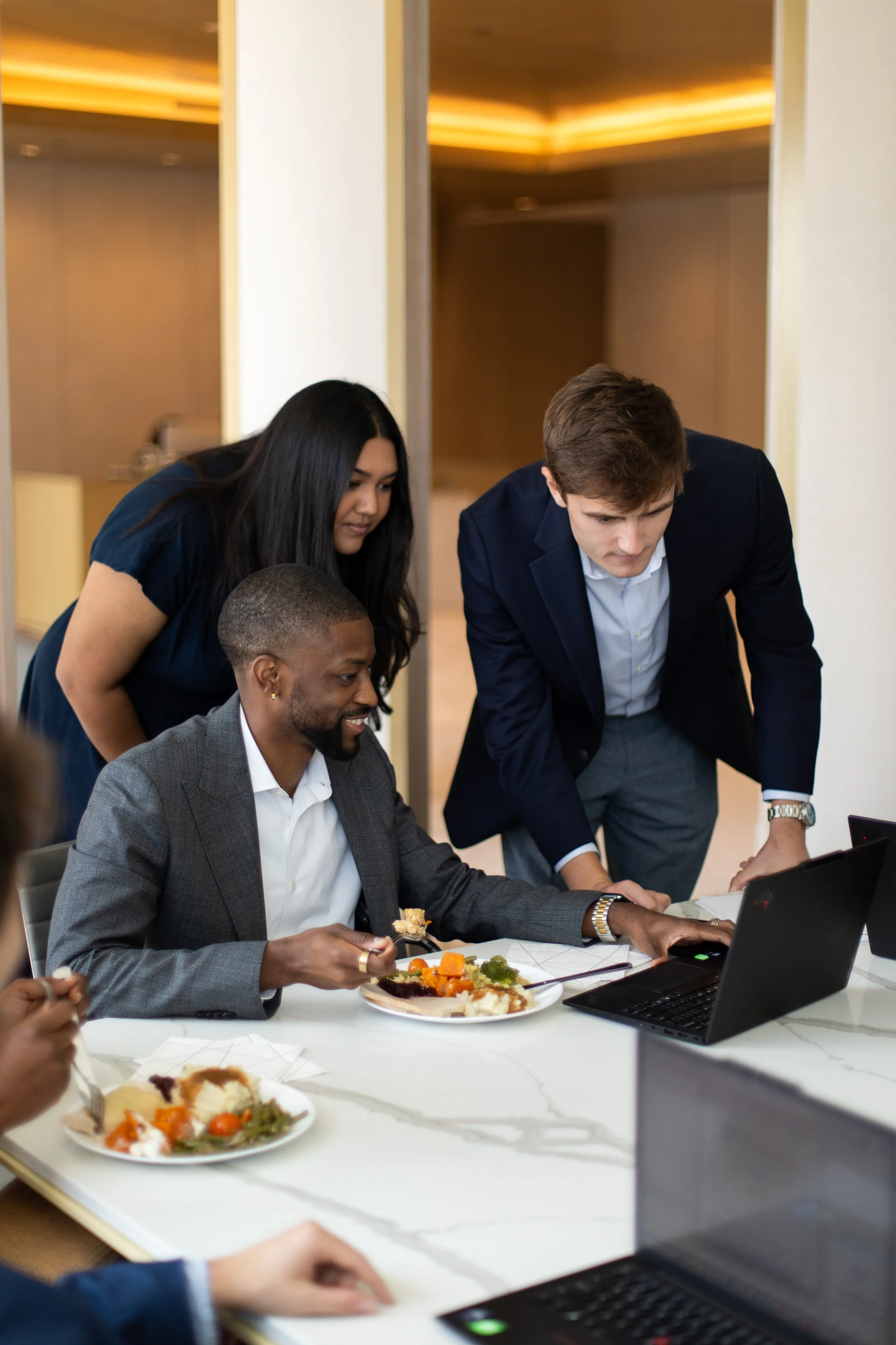 Three business professionals, two men and one woman, gathered around a laptop in a modern conference room. The man in a gray suit is seated eating food from a plate, while the woman with long dark hair and a navy top, and the man in a navy blazer are standing and leaning over, looking at the laptop.
