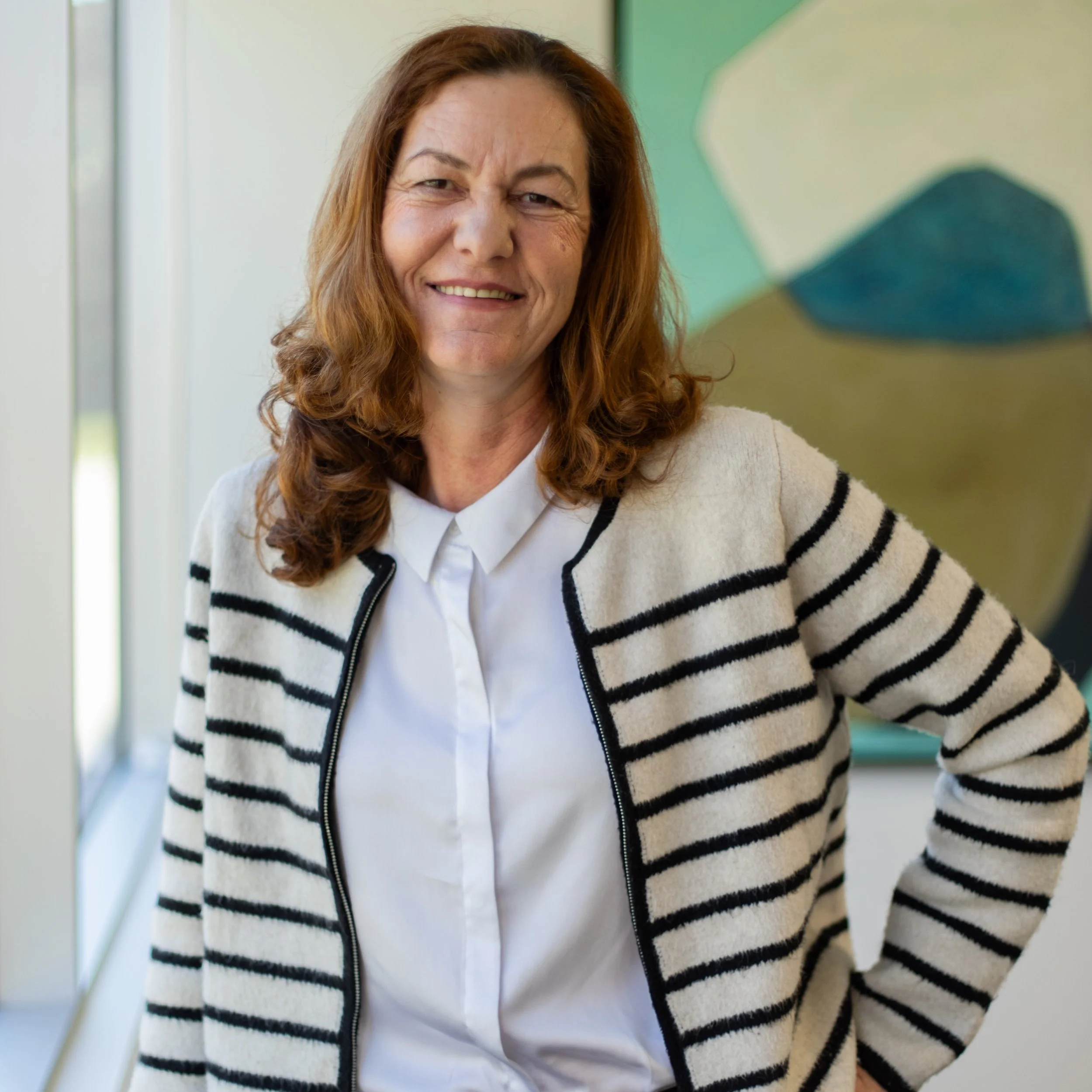 A smiling woman with long, curly, auburn hair wearing a white jacket with black stripes over a white shirt, standing indoors near a window with a blurred colorful background.