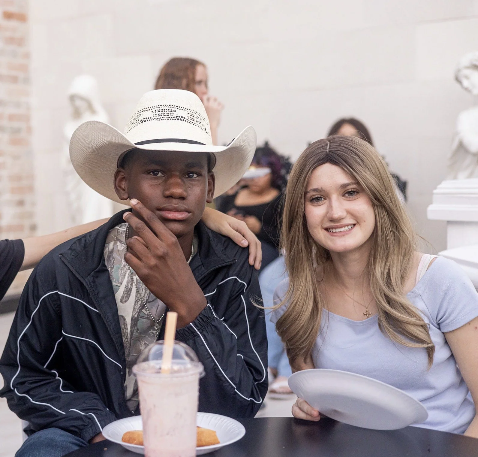 A young man wearing a cowboy hat and a young woman with long, wavy hair sitting together at a table in a cafe or restaurant. The man is touching his chin with one hand, and the woman is smiling and holding a plate. There are drinks and food on the table, and other people are visible in the background, along with decorative white bust sculptures.