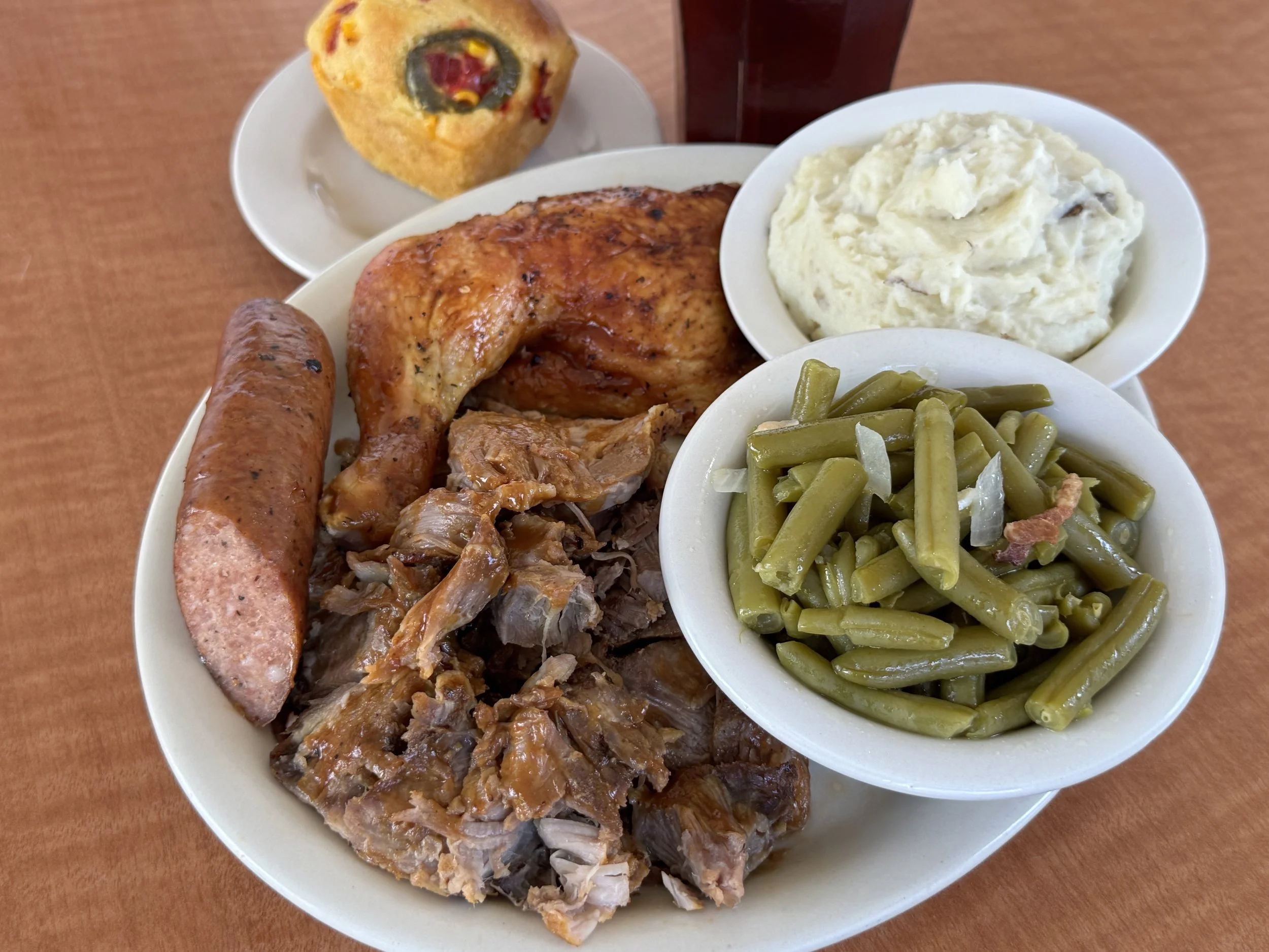 A plate of food with green beans, mashed potatoes, a baked chicken leg quarter, sliced turkey, sausage link, and a side of stuffing. There is also a slice of cornbread and a glass of a dark beverage.