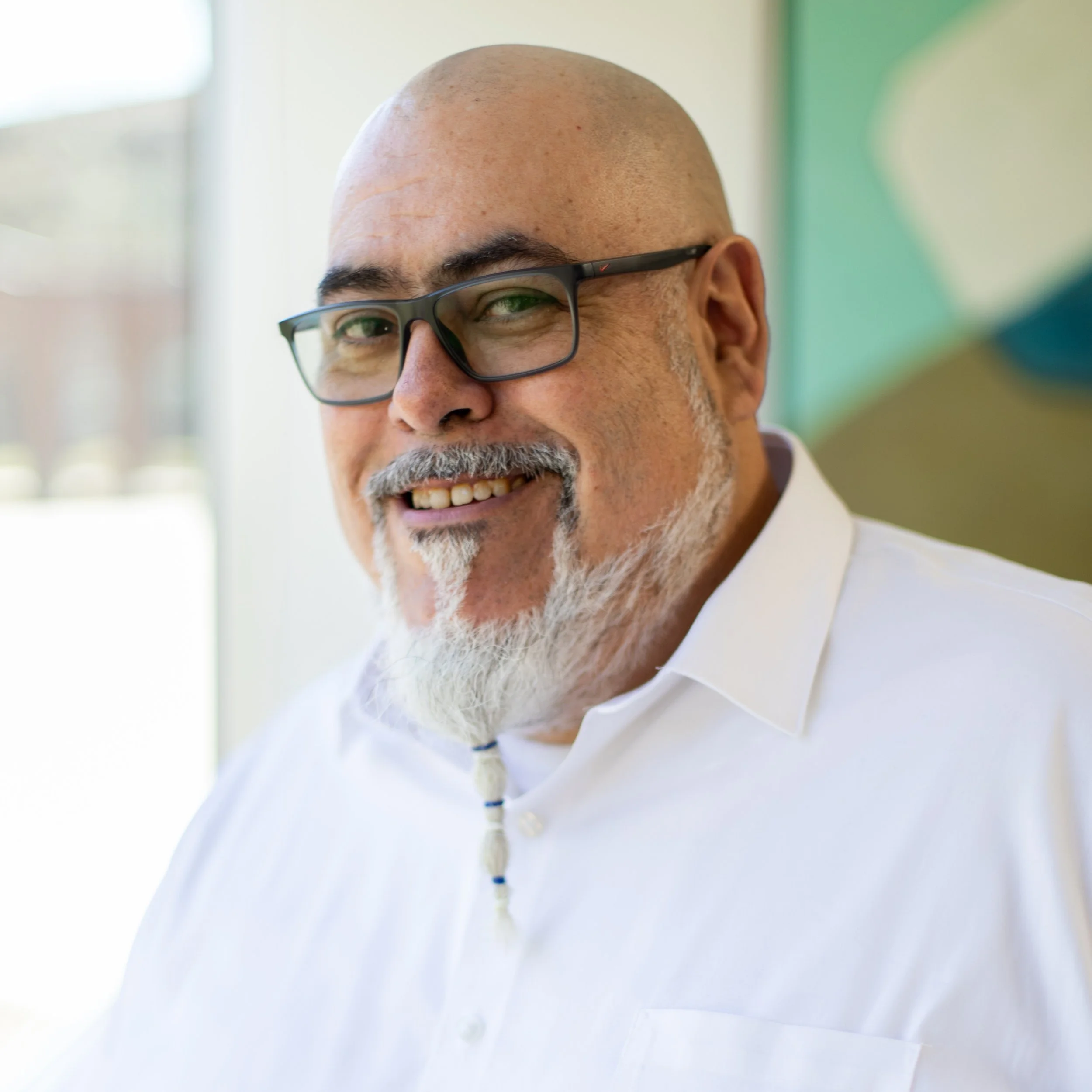 Close-up of a smiling man with a bald head and glasses, wearing a white shirt, indoors with a blurry background.