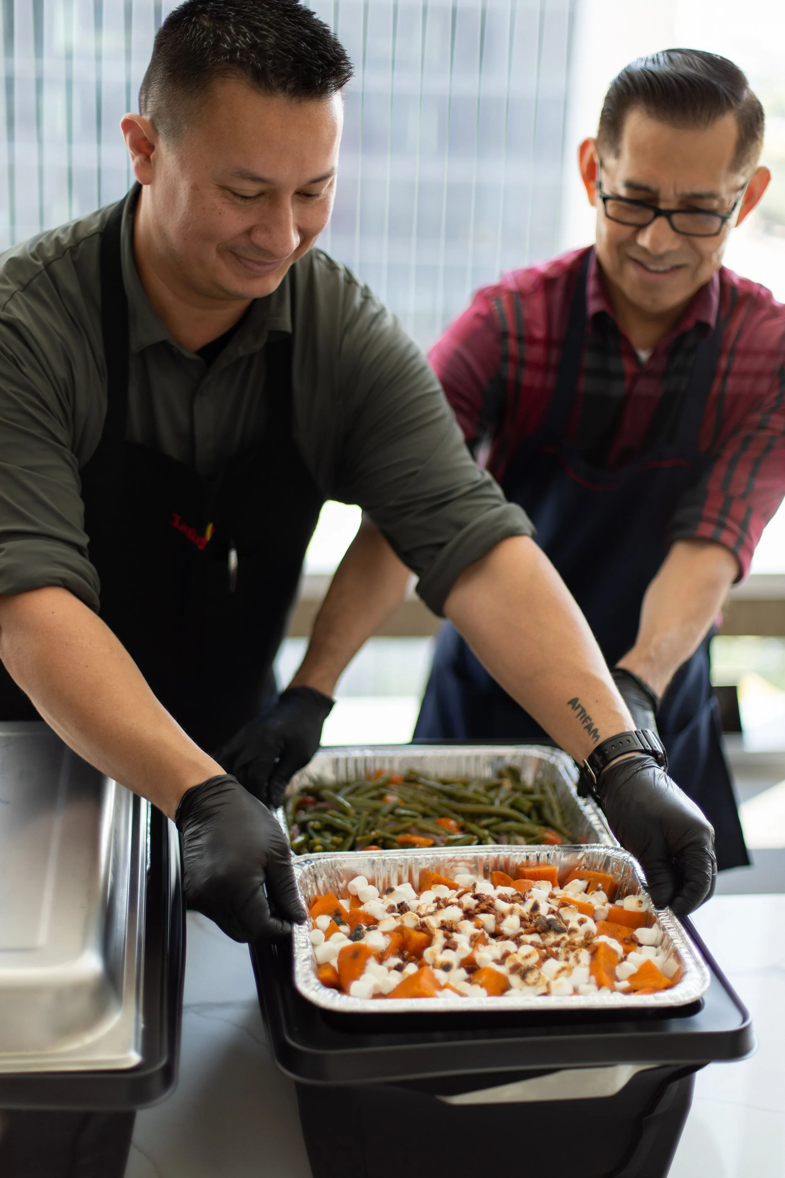 Two men wearing black gloves and aprons preparing trays of food, including sweet potatoes topped with marshmallows, green beans, and carrots, in a well-lit indoor space with large windows.