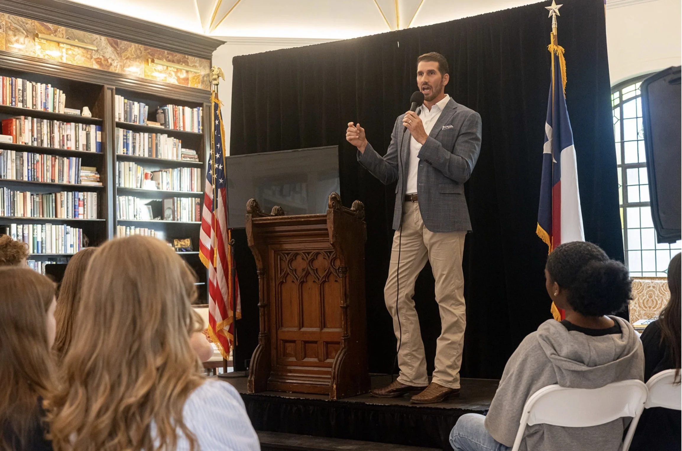 A man in a gray blazer and beige pants is speaking into a microphone while standing at a wooden podium during a presentation or speech. He is in a room with bookshelves, American and possibly Texas flags, and a large window with stained glass. The audience of young people is seated and listening attentively.