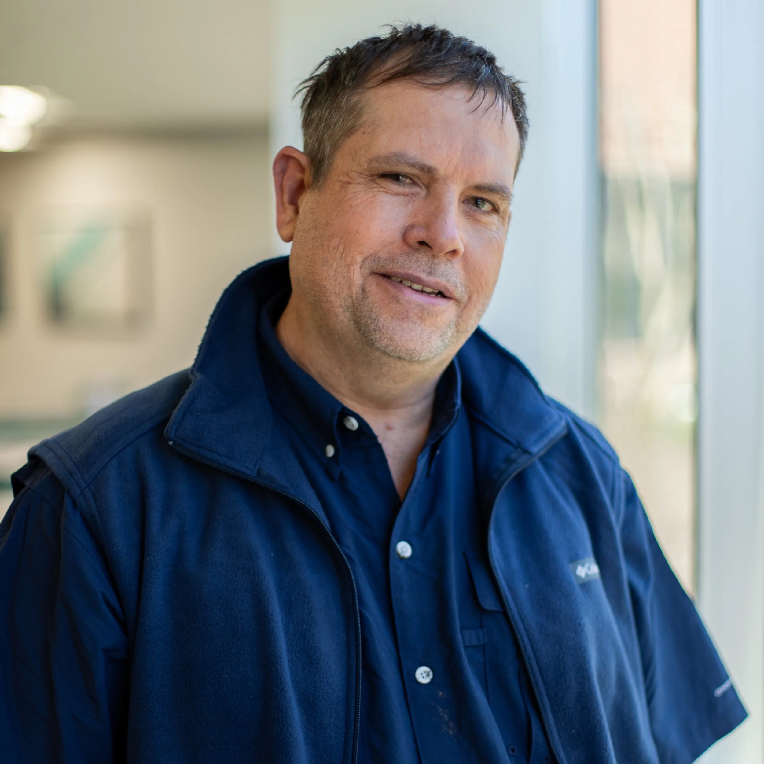 Close-up of a middle-aged man with short dark hair, light-colored eyes, wearing a dark blue jacket and a collared shirt, smiling, standing indoors near a window with soft natural light.