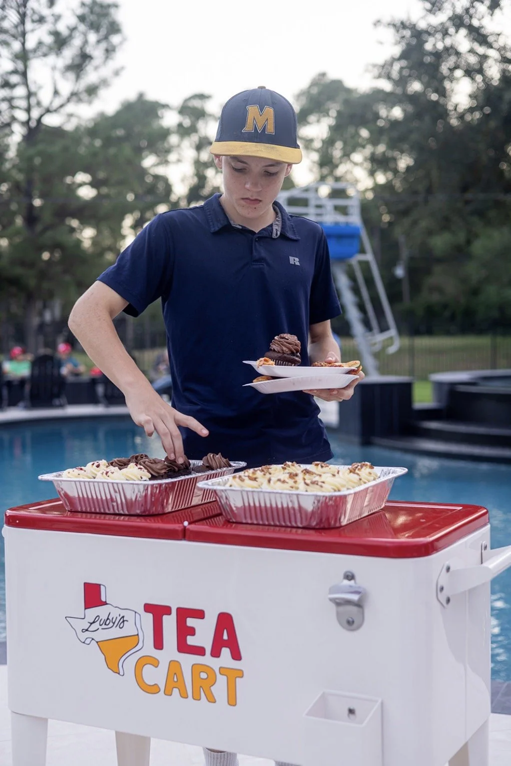 A young person wearing a baseball cap and navy-blue polo shirt serves brownies on a white plate from a white and red tea cart near a swimming pool.