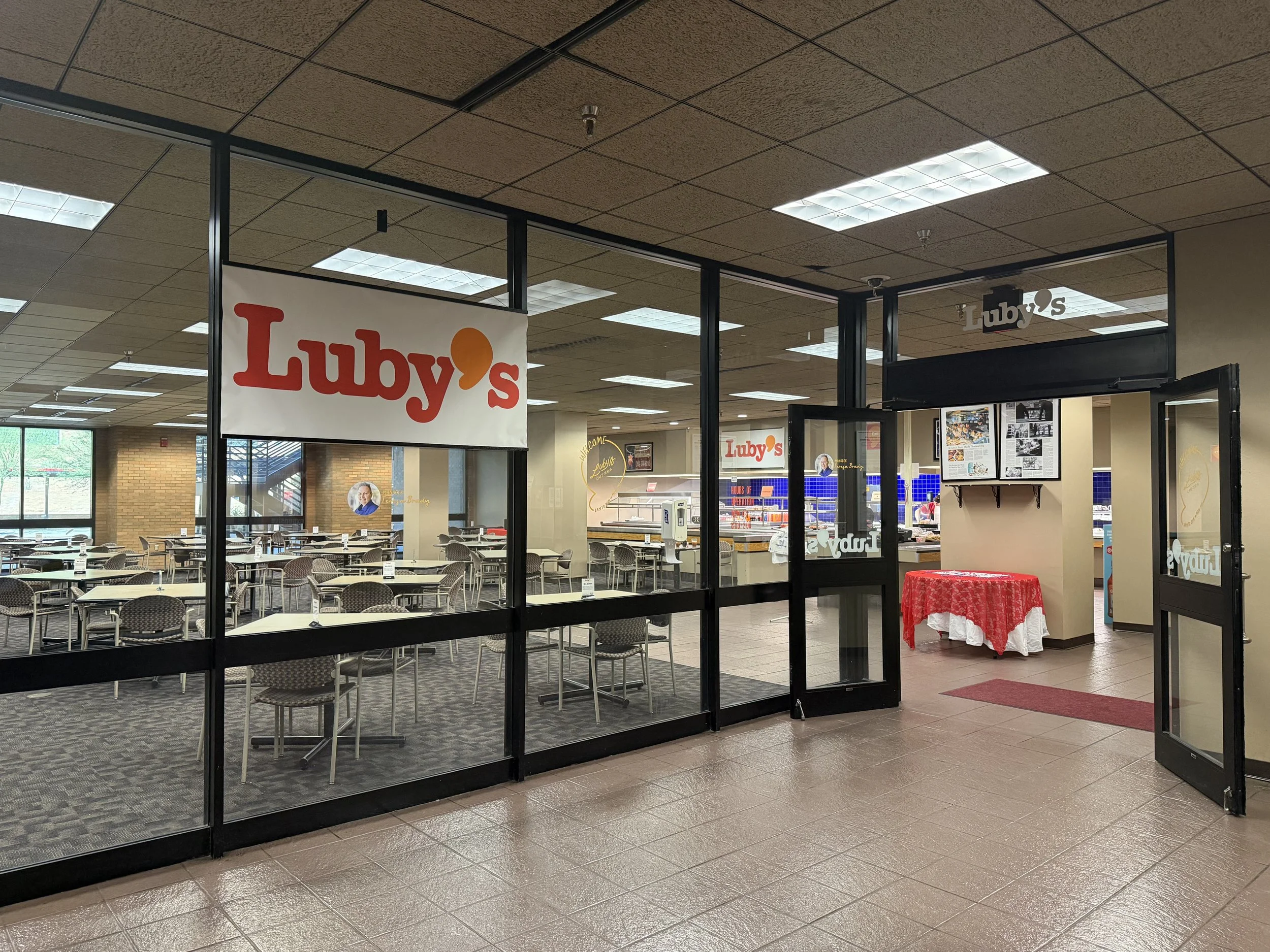Inside a restaurant or cafe named Luby's, with several tables and chairs, and a red tablecloth-covered table near the entrance.